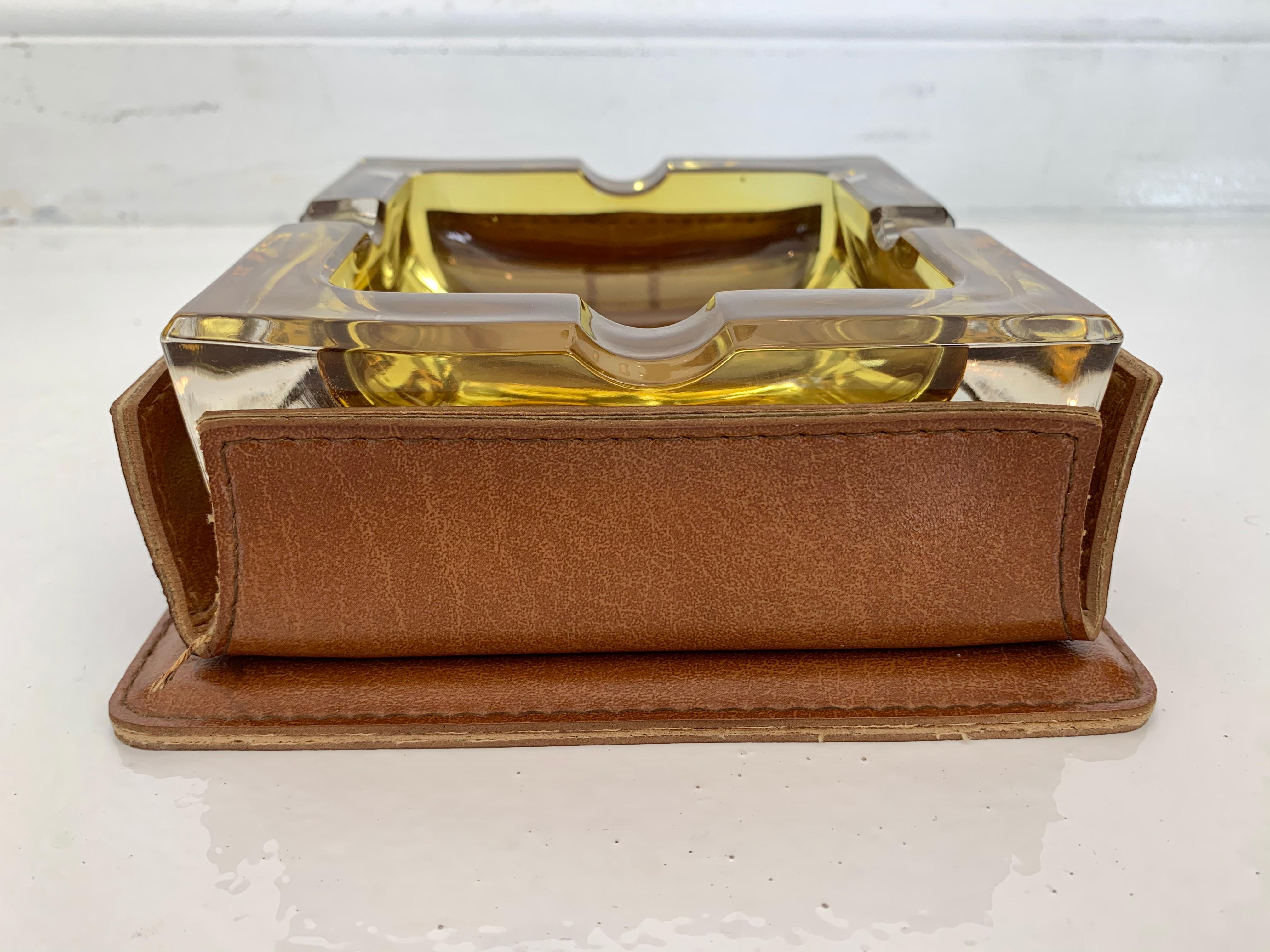 Handsome saddle leather and glass ashtray/ catchall in the style of Jacques Adnet. Square saddle leather holder with contrast stitching. Amber glass dish can be removed for cleaning. Good condition. Great desktop object.