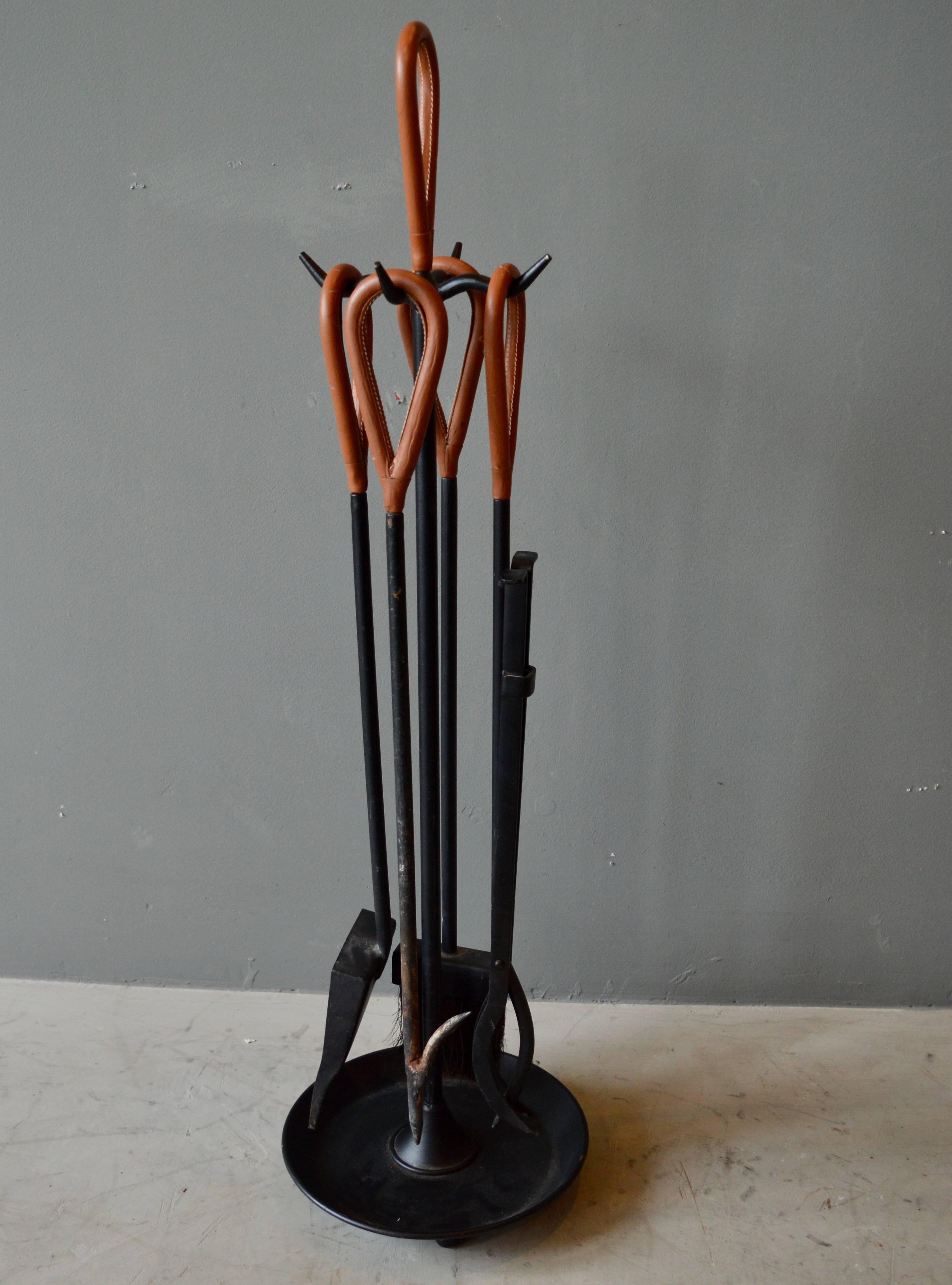 Gorgeous fireplace set in the style of Jacques Adnet. Iron frame with saddle leather wrapped handles. Fireplace set includes Stand along with four tools, broom, dustpan, tongs and fire poker. Very good vintage condition.