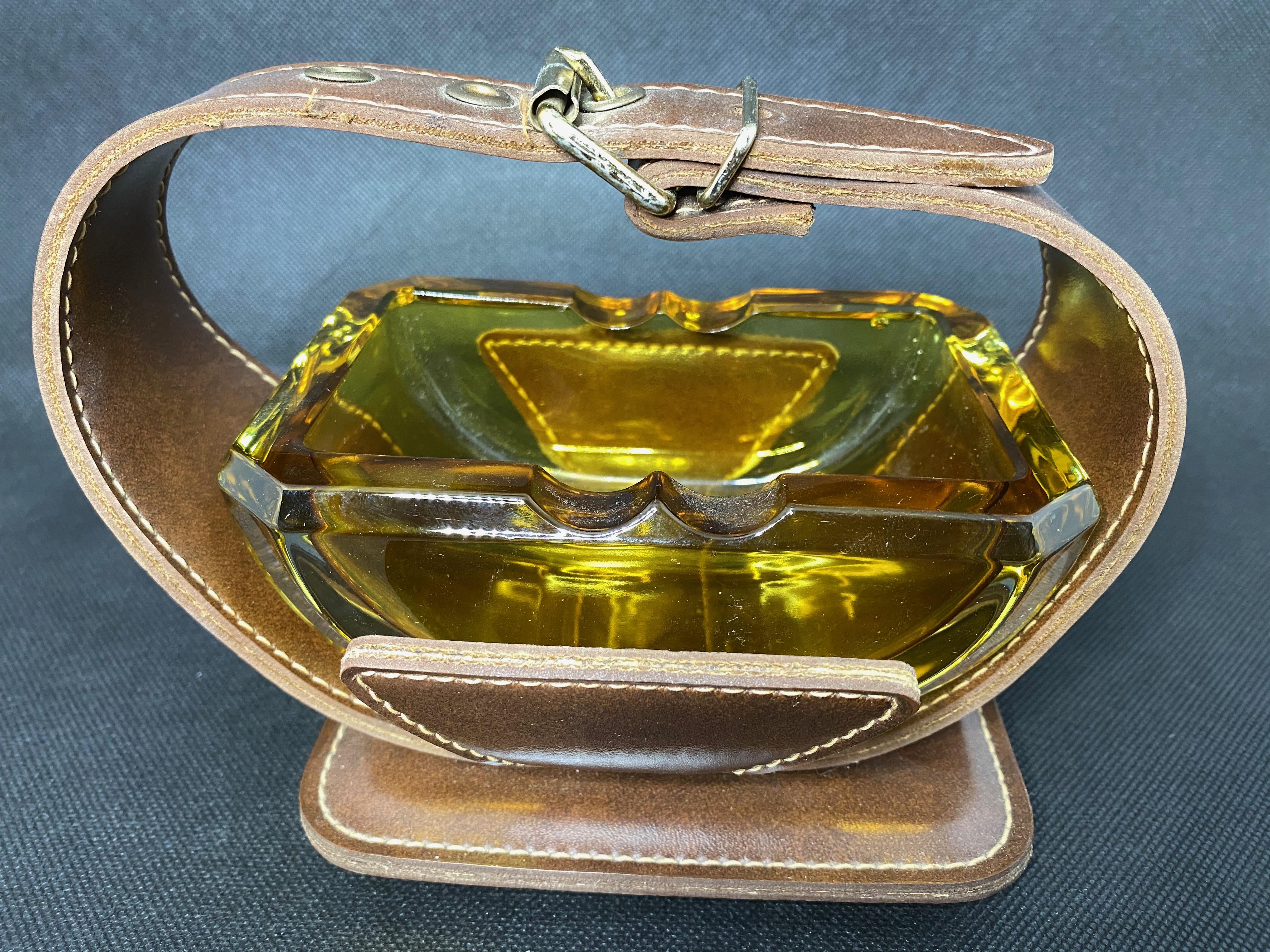 Leatherette, brass and glass ashtray in the style of Jacques Adnet. Leatherette frame with brass studs. Inset amber glass dish. Great tabletop object. Great vintage condition.