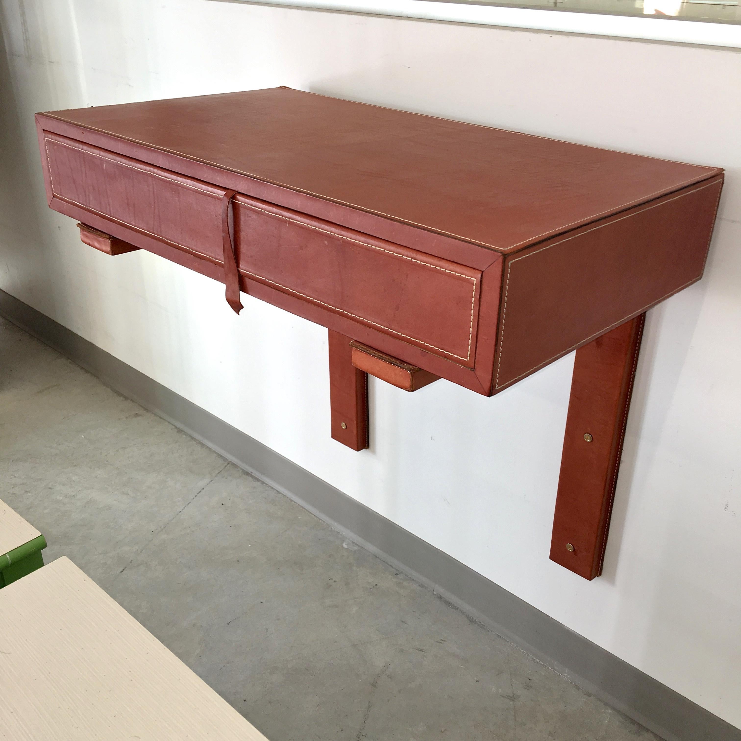 A wall console wrapped in saddle leather supported by two L shaped brackets with a top stitch detail in the manner of Jacques Adnet. One drawer with a piece of leather serving as the pull. Would serve well as a desk, or sideboard.

