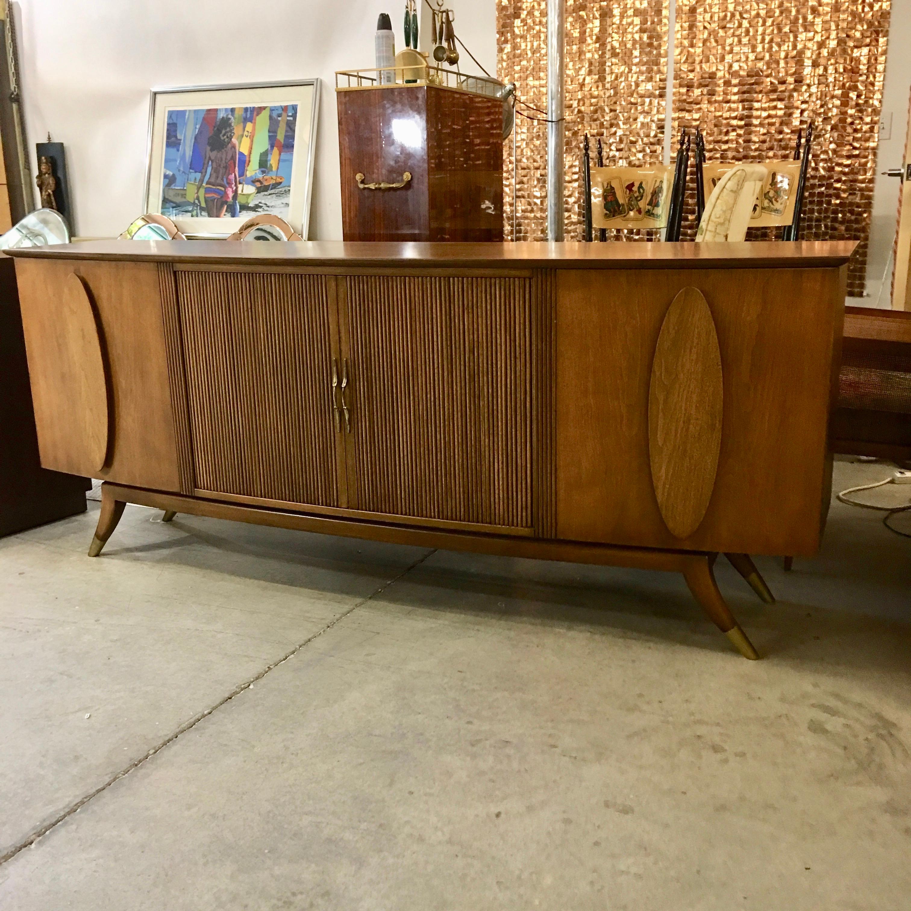 Long bow-fronted credenza in walnut with two cabinet doors and four drawers behind sliding tambour doors with brass pulls. The top two drawers are lined and divided for silverware. The two side cabinets each have two horizontal adjustable shelves.