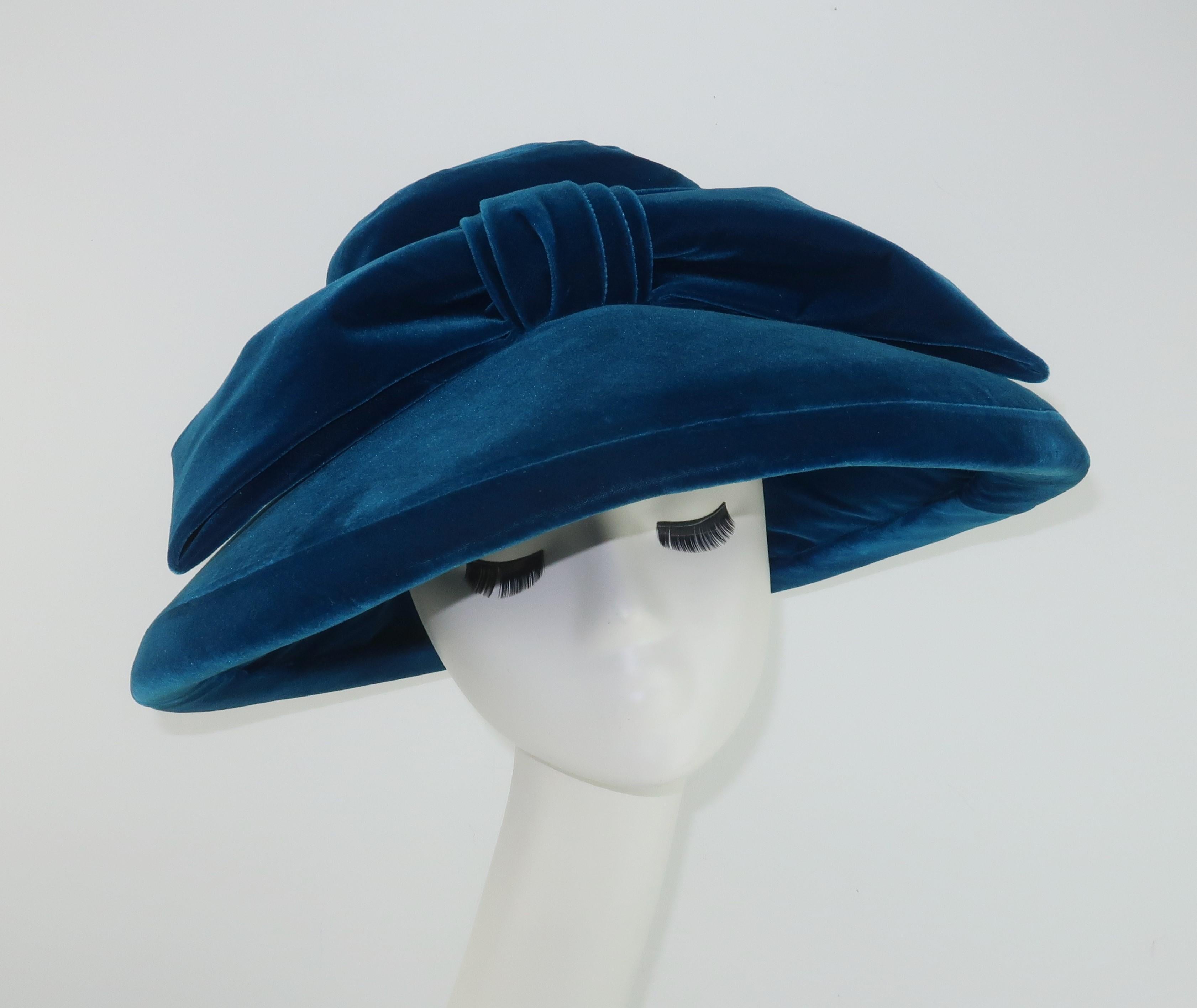 A wide brimmed velvet hat by Adolfo in a lush peacock or teal blue.  Festooned at the band with a large bow and designed with a mushroom style shape both framing the face and also allowing for an incognito look when worn with large shades ... a la