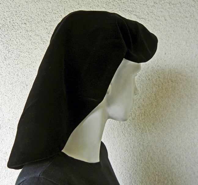 Uno snood in morbido velluto nero di Adrian Original che simula un cappuccio da suora.  Sostenuto da un cerchio esterno.  Sia lo snood che il cerchio sono rivestiti di rete.  Un design molto semplice ma brillante con una silhouette forte.  Forse