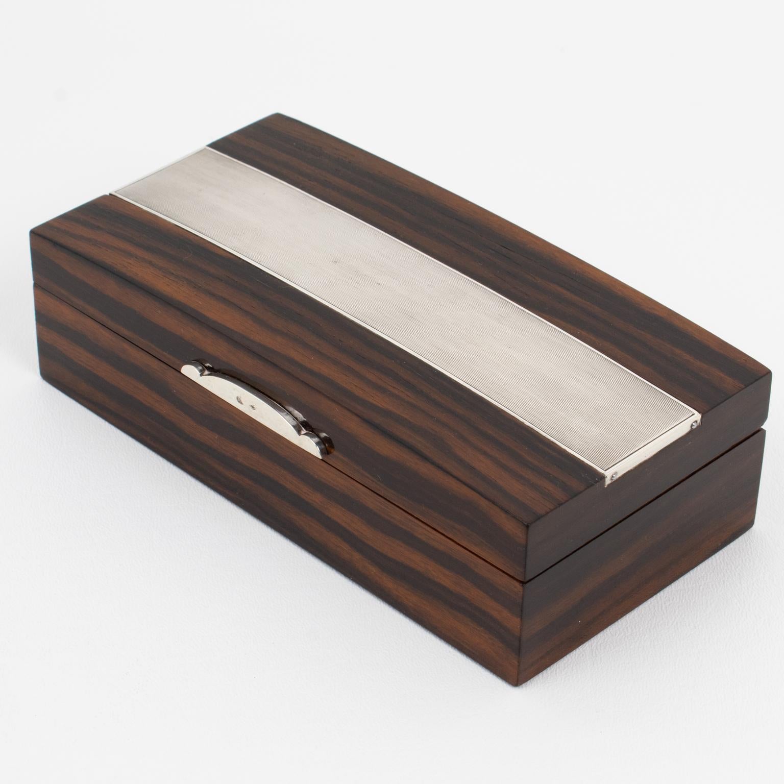 Se trata de una lujosa caja Art Déco de madera y plata de ley con tapa, hecha a mano por la orfebre Adriana Binazzi en Florencia (Italia) en la década de 1930. La forma rectangular ostenta una composición refinada con un cuerpo de madera de Macassar