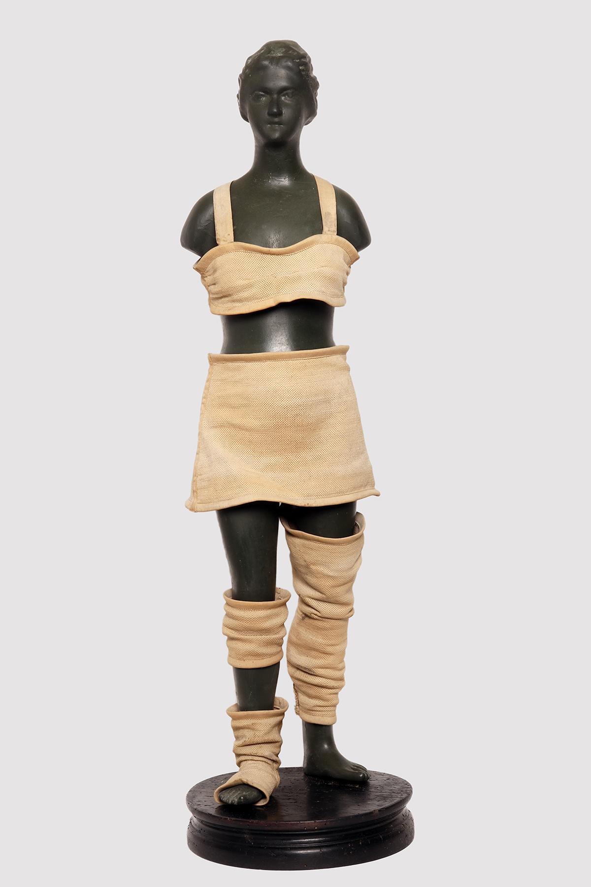 Advertising mannequin for elastic and containment belts. The base is made of wood. The mannequin is made of uniform black buffered papier-mâché. The mannequin is intended to demonstrate the use of orthopedic aids by advertising it. It shows various