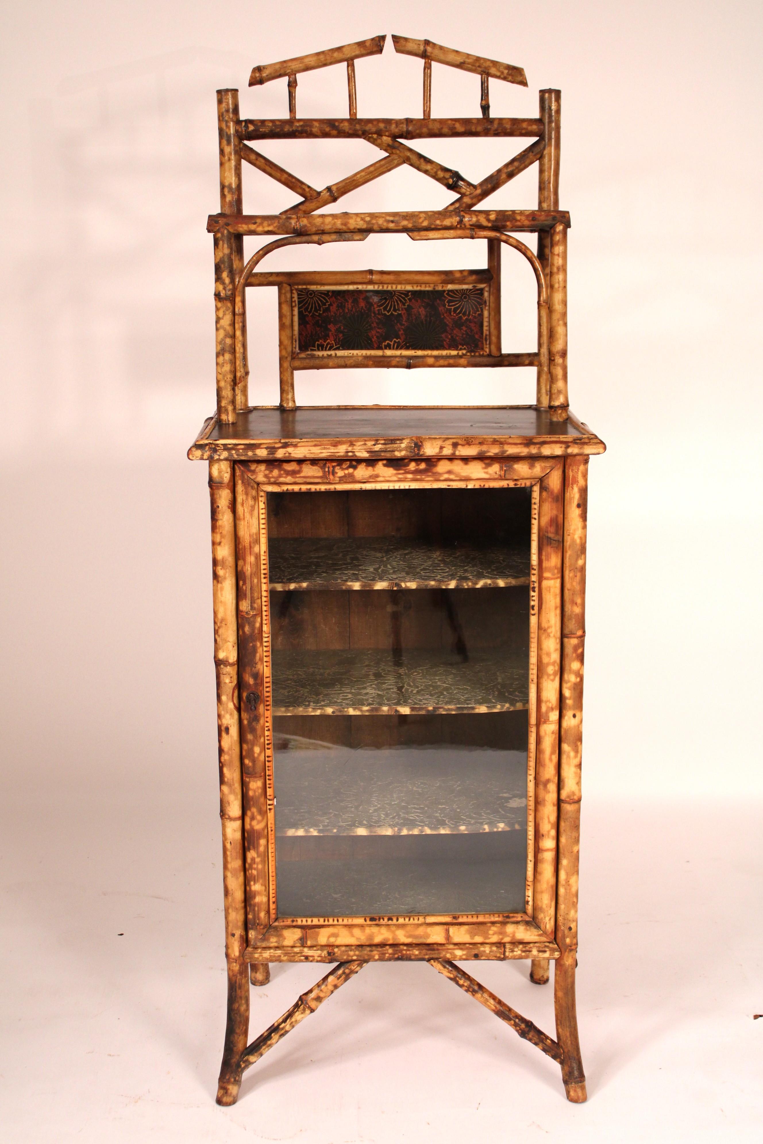 With a bamboo pediment with an inset lacquer panel, lacquered top, raffia sides, a bamboo framed glass door with 3 shelves, resting on bamboo legs.