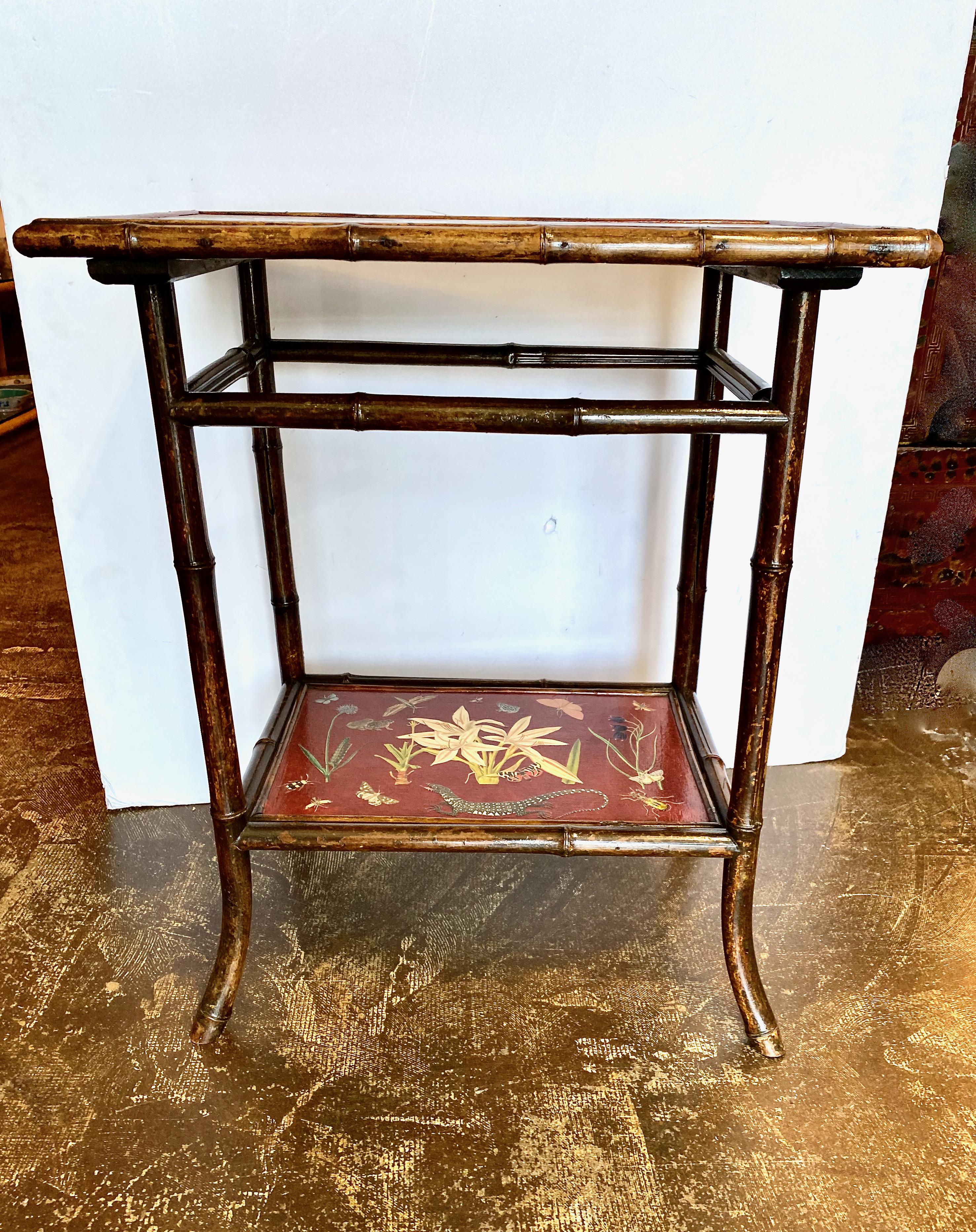This is a charming early 20th century English Aesthetic Movement Side or Occasional Table. The table features a burnt bamboo frame supporting two tiers of beautifully shelves. The brick red base surface is decorated with vintage colored print cutout