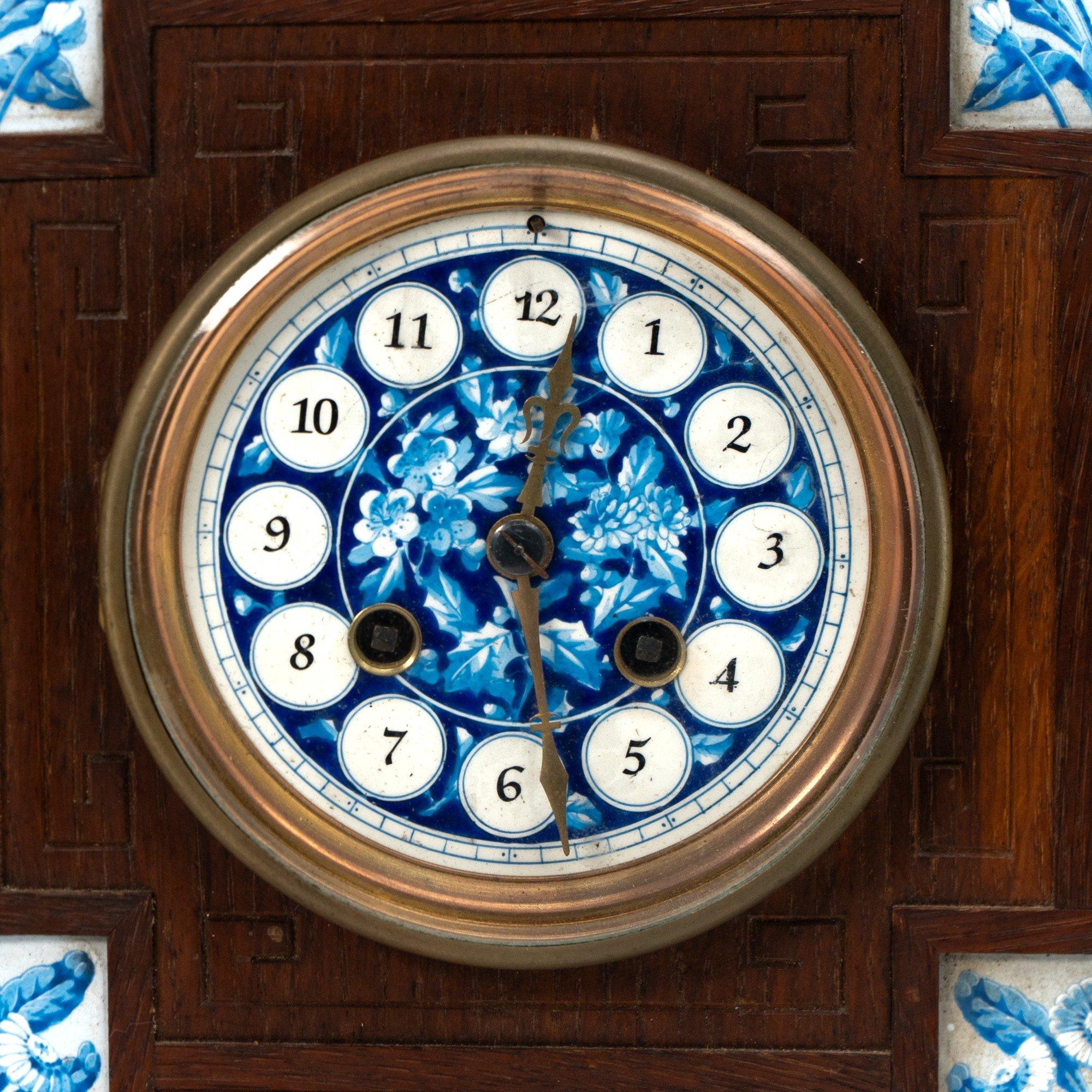 Howell & James, in the style of Lewis Foreman Day. An Aesthetic Movement oak mantle clock with a carved cresting to the top and fan-shaped detail, framed by incised decoration and a shaped apron to the base.
The circular dial is decorated in blue