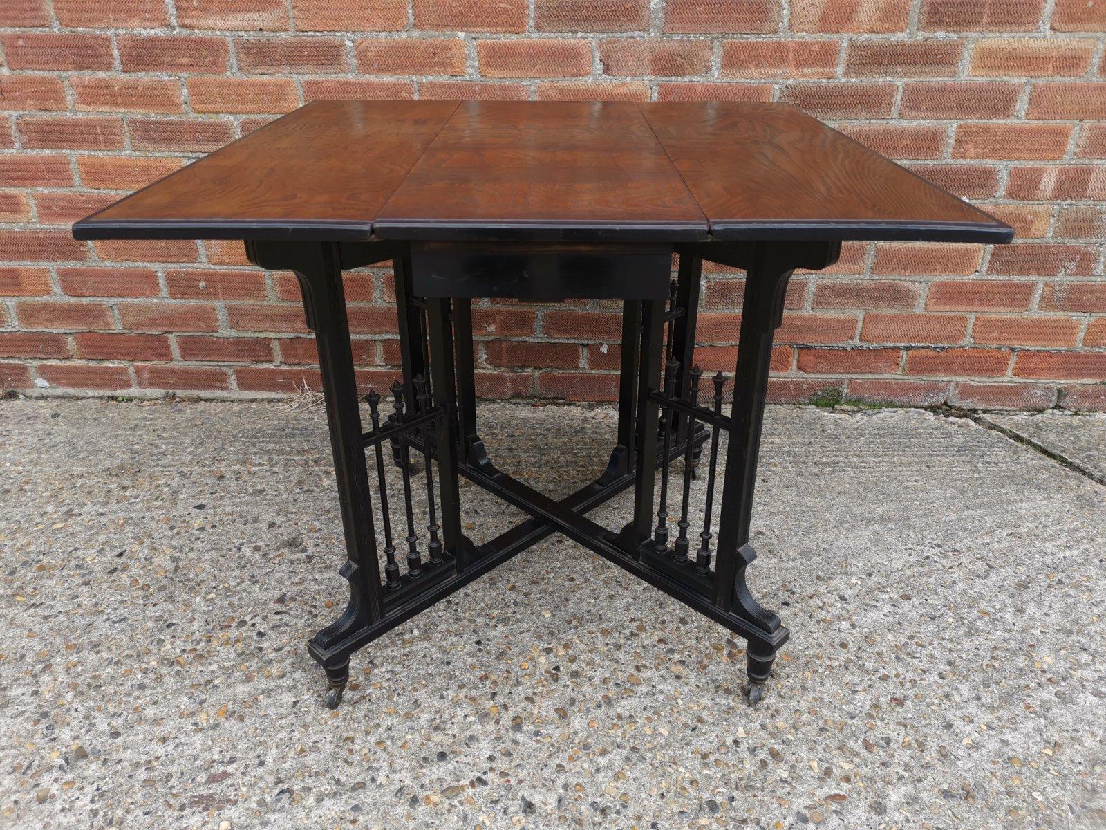 A Continental Aesthetic Movement scissor action drop leaf side table with a wild grain Hungarian Ash top. A secret drawer that has a button underneath to open it. An ebonized undercarriage with elongated turned spindles and tramline incised details