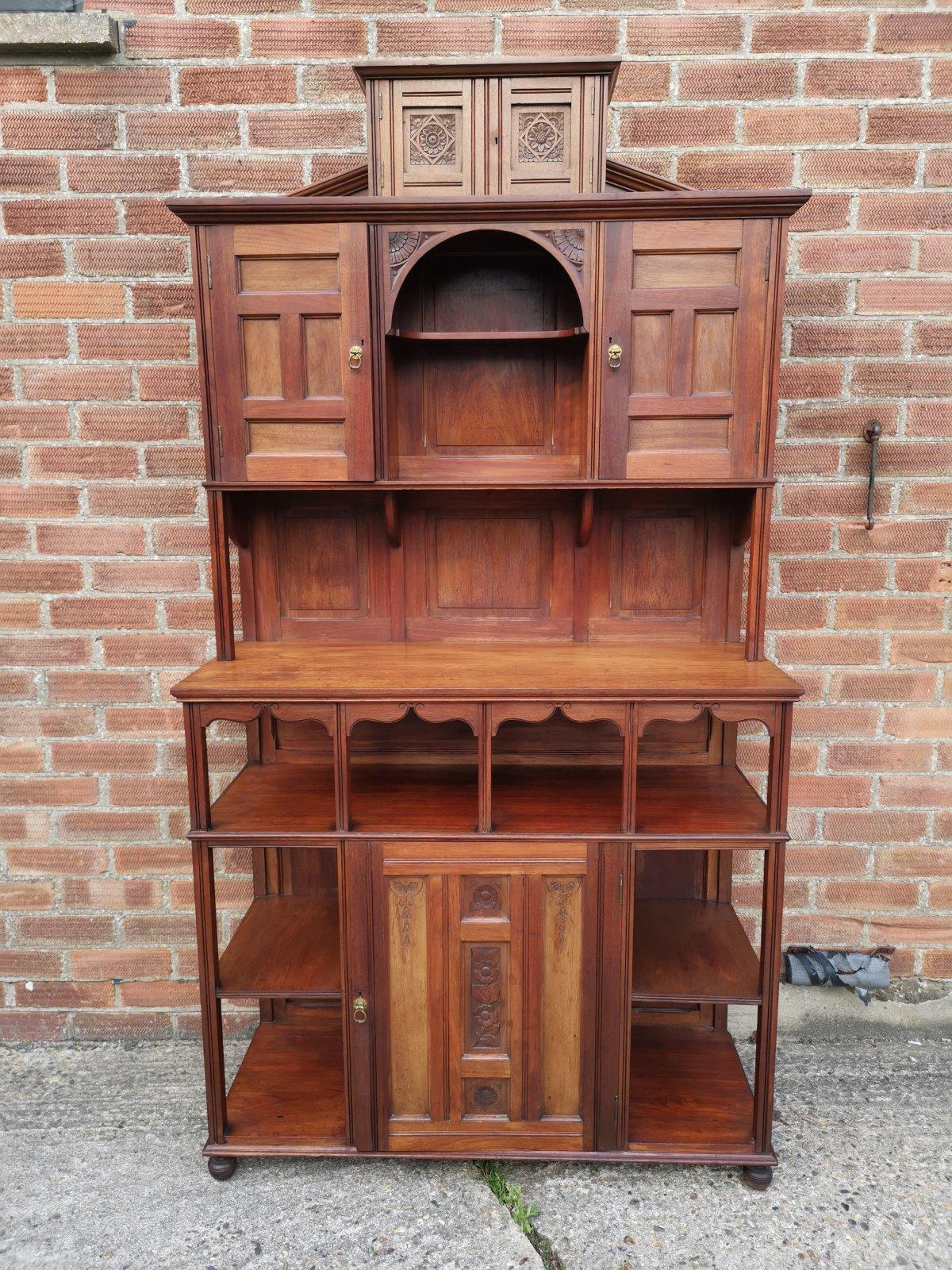 An English Aesthetic Movement walnut cabinet of architectural form with carved floral decoration to the upper and lower cupboard doors with stylized brass ring pull handles to the doors, carved quarter sunflower lunettes to the upper section and