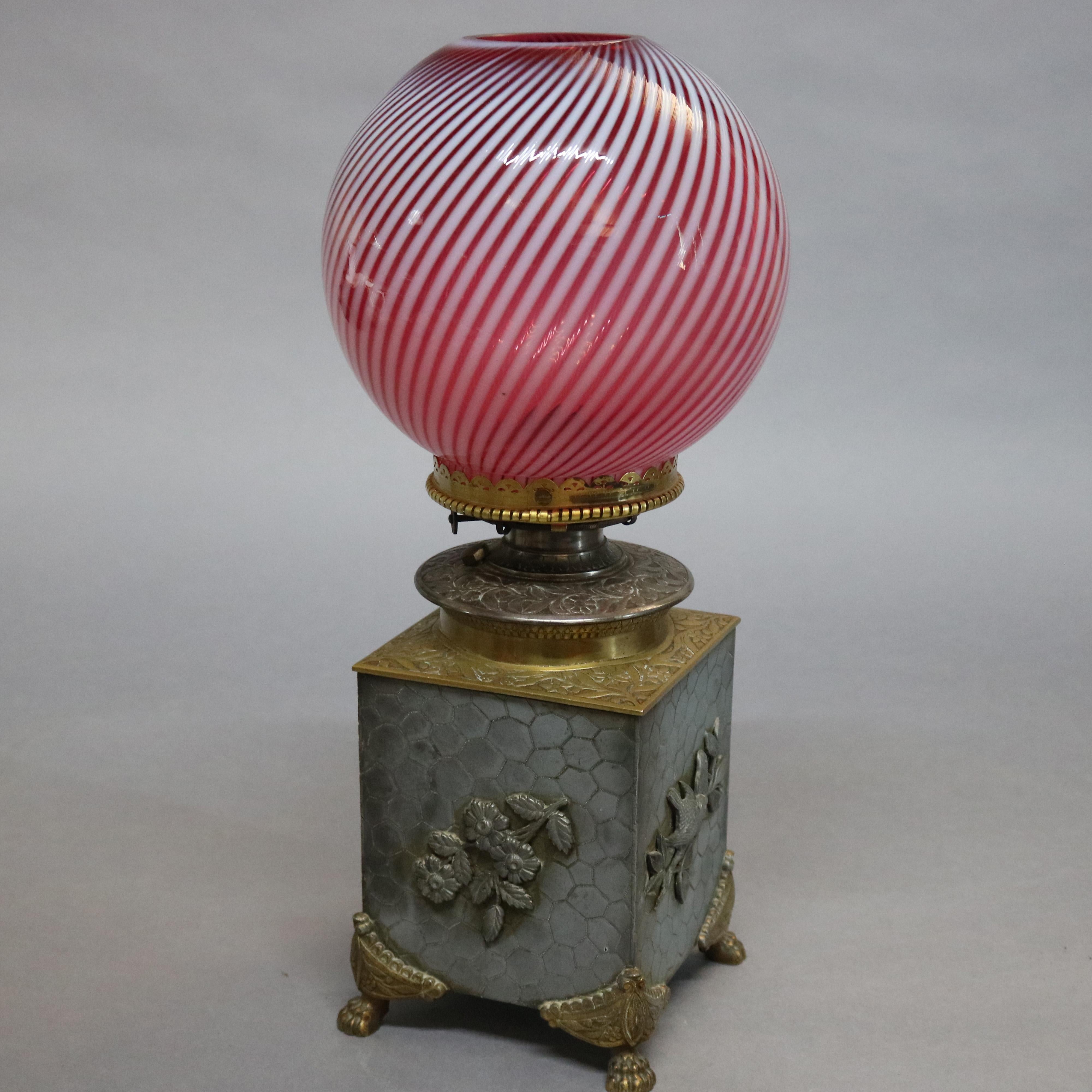 An antique Aesthetic movement oil lamp offers cube form base with honeycomb allover design having cast foliate reserves, gilt foliate decorated cap and raised on gilt paw feet, cranberry swirl glass shade surmounts, circa 1870.

Measures: 17.5