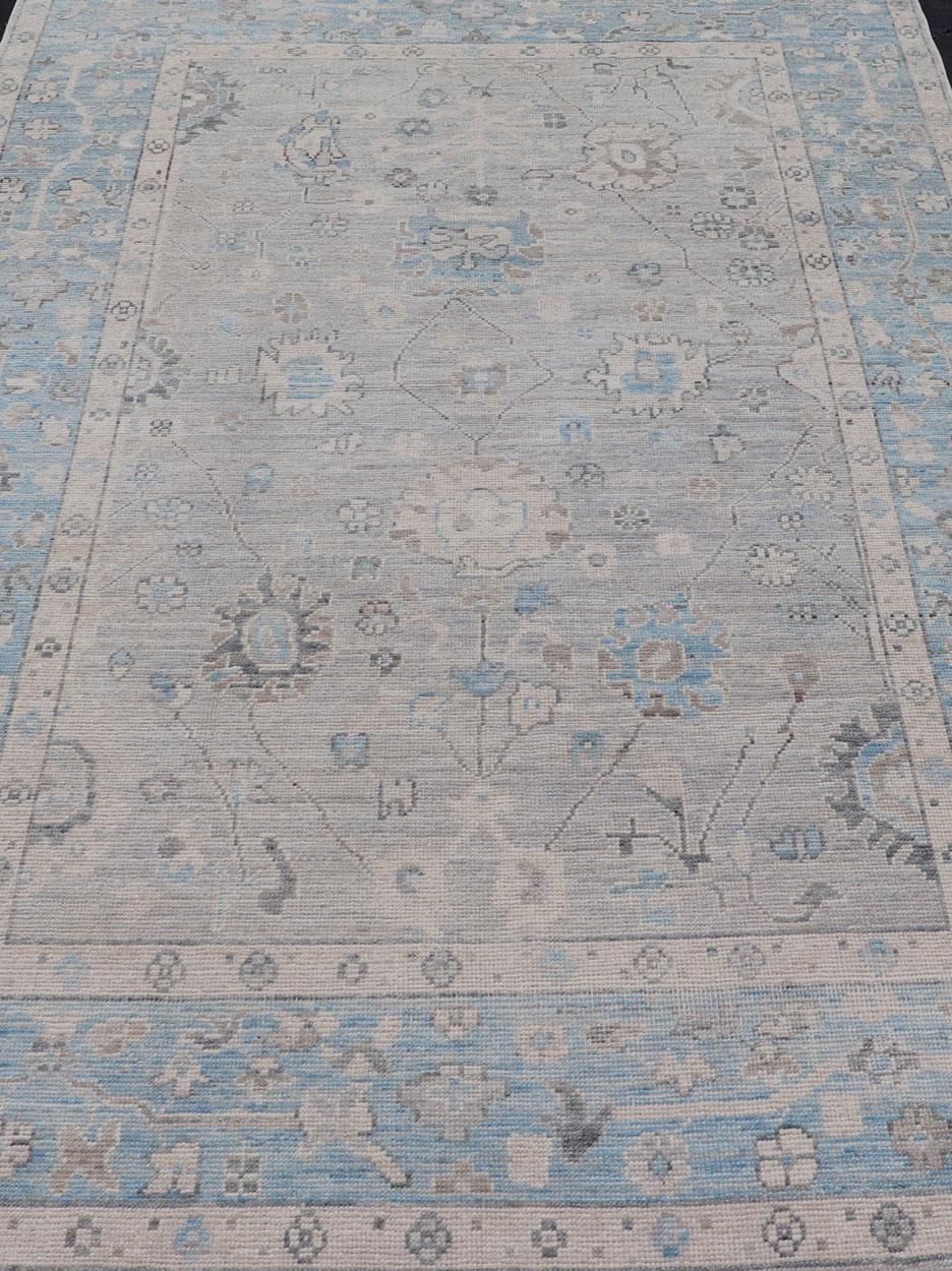 Nouée à la main en Afghanistan, cette pièce rustique est charmante avec sa bordure bleu poudre et son fond gris clair. Au-dessus se trouve un modeste motif floral de style Oushak s'étendant sur toute la bordure et le champ. Le design discret et les