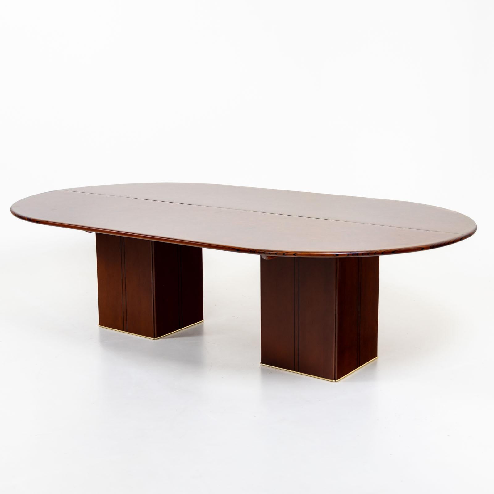 Large dining table or conference table with oval tabletop and veneer of walnut and birch burl. The table rests on two square legs, which are stabilized by means of concrete counterweights. The weights are embossed with the logo of Maxalto as well as