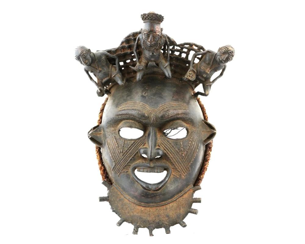 An African bronze mask from Benin. Featuring wide-open eyes, a gaping mouth with protruding teeth, and prominent nostrils. The face is intricately detailed with numerous tattoos. The neck is adorned with a semicircular plaque, while three small