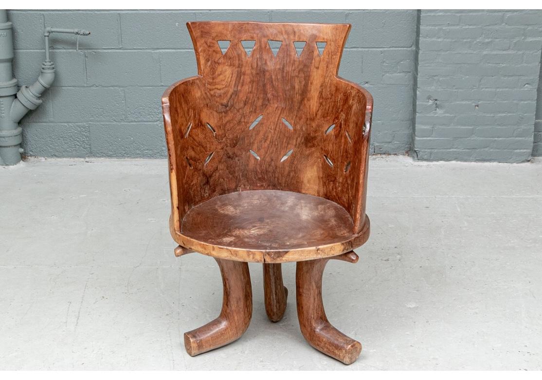 A heavy carved natural hardwood arm chair with high back and cut out slots and triangle motifs. The tub mounted on a single circular slab for the seat. Raised on three heavy curved cylindrical legs with flat ends. The natural wood grain in different