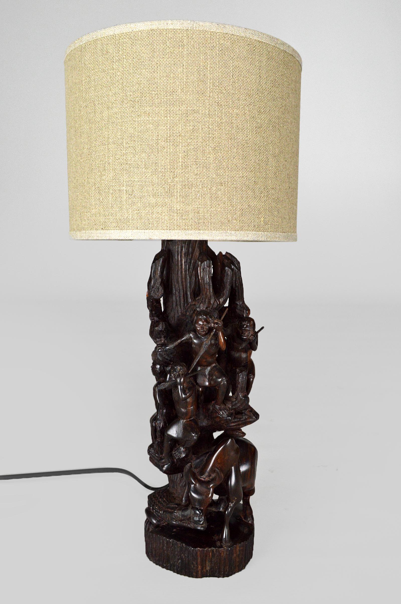 Rare African sculpture mounted as a table lamp.
Ebony base, linen lampshade.

Handmade sculpture in a single block of wood representing a group of 5 hunters/warriors with spears, taking refuge in a tree, above a buffalo attacked by a