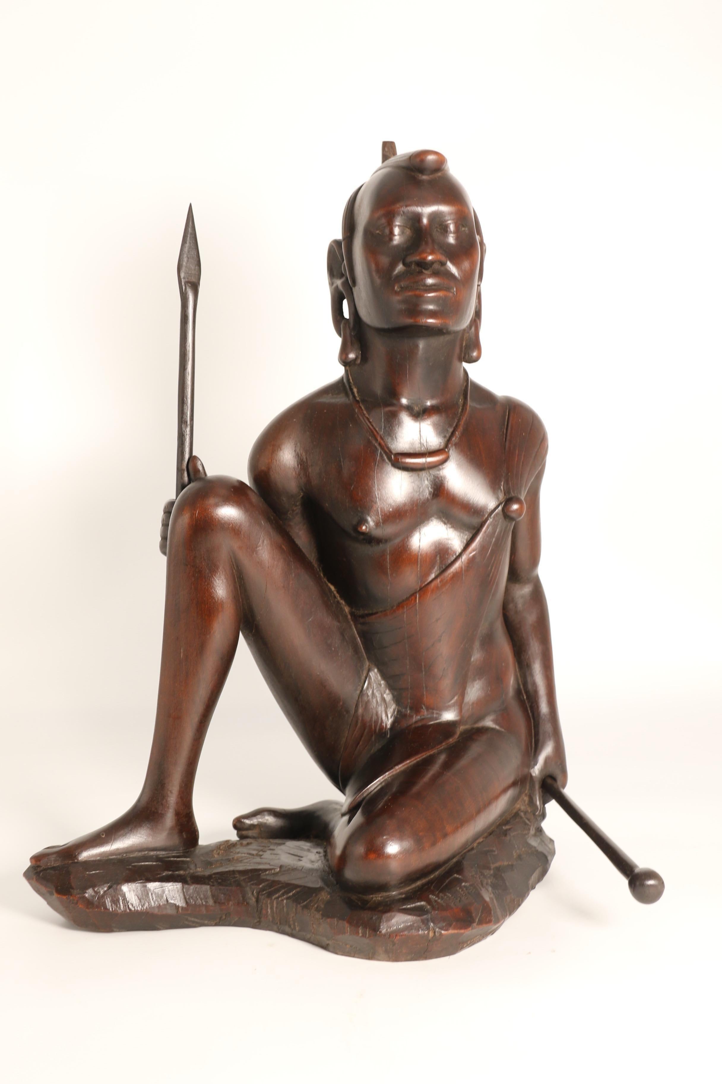 Questa splendida scultura in legno massiccio intagliato rappresenta un guerriero africano Maasai seduto in posizione semi-inginocchiata. An He ha lineamenti maschili e potenti e capelli acconciati in modo impeccabile. An He è semi-nudo con un fisico