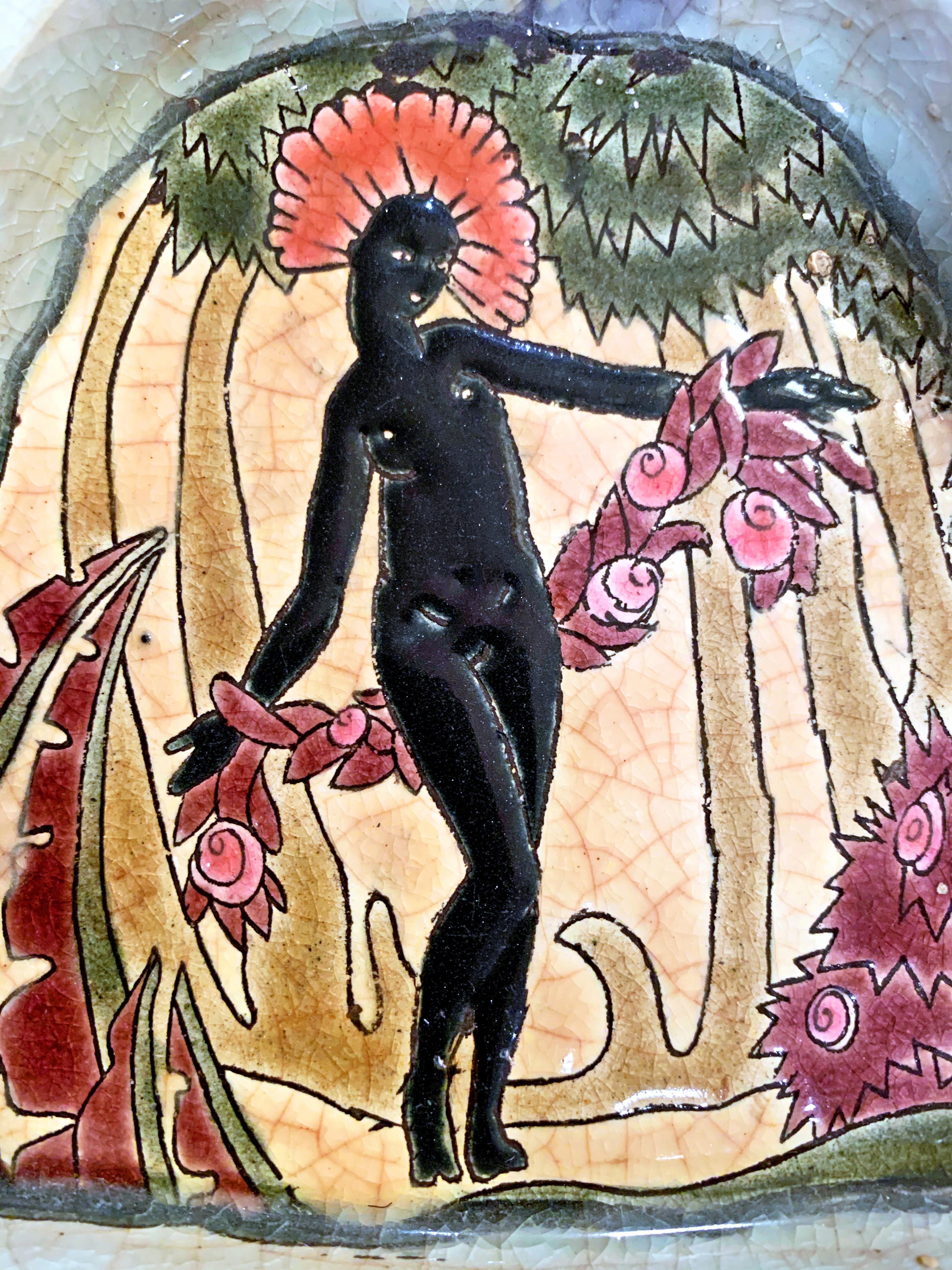 Brillamment émaillé dans des tons de corail, de noir de jais, de prune et de mousse, ce très rare plateau de courtoisie représente une figure féminine africaine avec une coiffe de plumes et des guirlandes de fleurs, dans un décor tropical encadré de