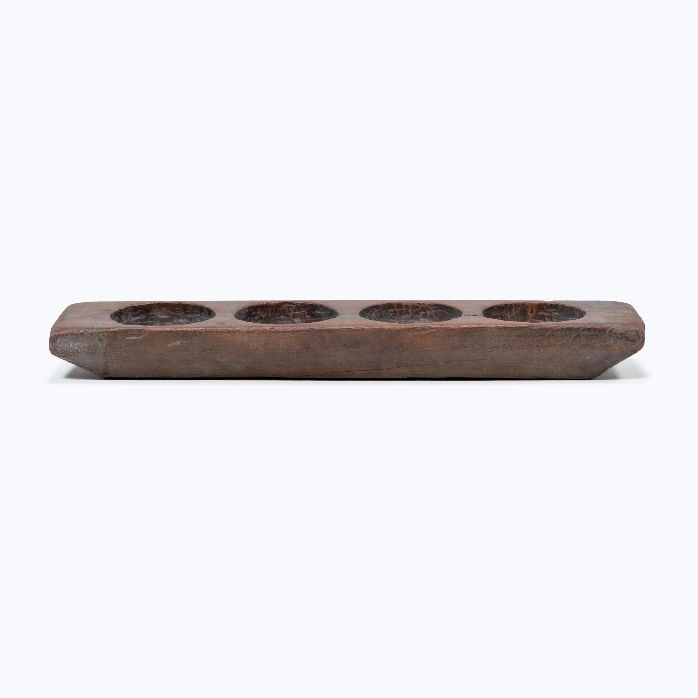 This unusual rectangular tray was originally used as a mortar for grinding and removing the husks of cereal grains. The multiple recesses provided a convenient way to prepare and store multiple grains and spices for a meal. The tray's trough-like