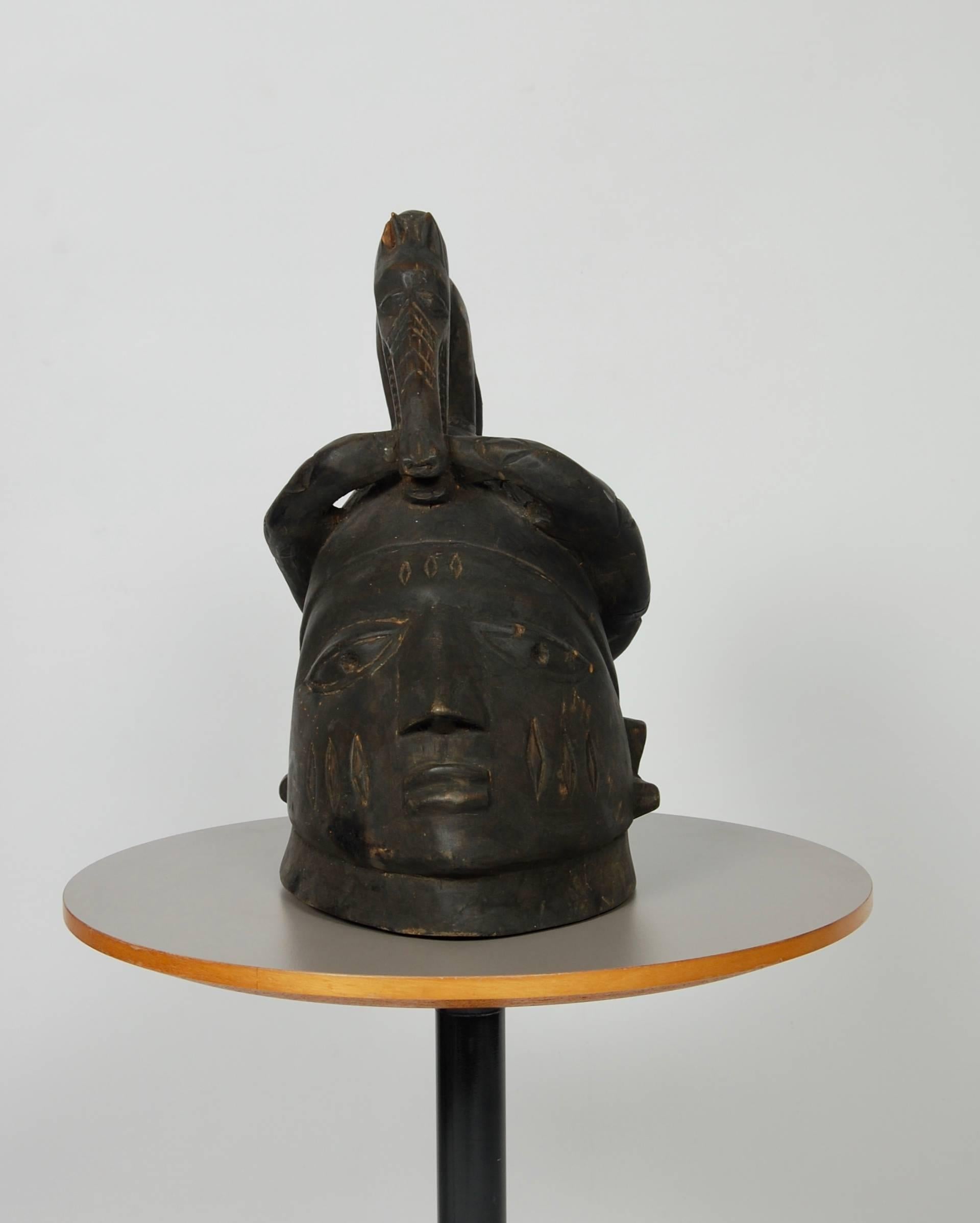 Blacked wood helmet mask (Gelede), large human head surmounted by a dog eating a snake which curves around the head. No perforation for attachment to costume from Yoruba, Nigeria. Purchased in 1982 at James R Lawson Pty. Limited, Sydney, Australia