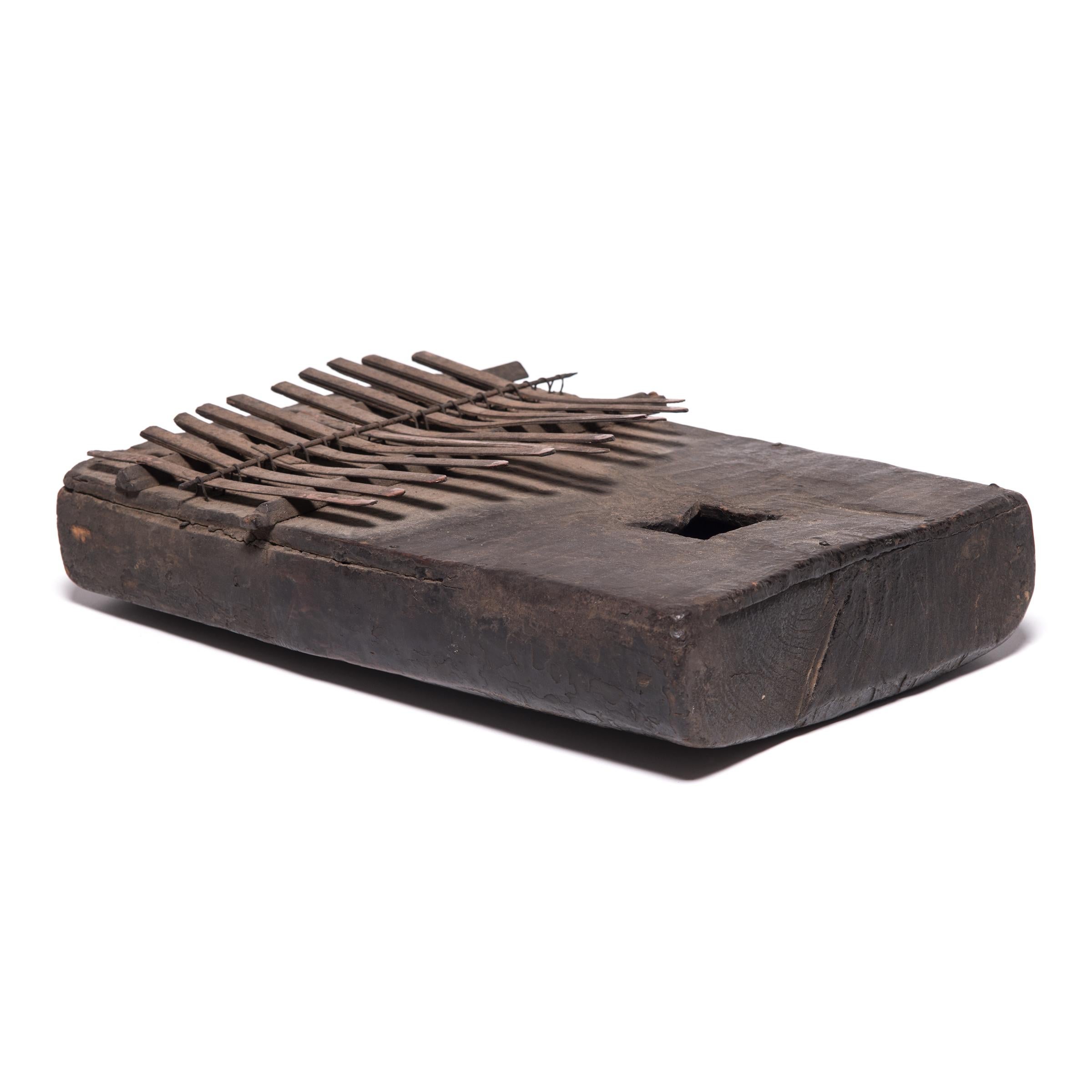 This rectangular object is a thumb piano from Central Africa, commonly known as a mbira, sanza or kalimba. The instrument is comprised of a resonance chamber with a sounding hole and a set of twelve keys, formed of a hollowed-out wood board and thin