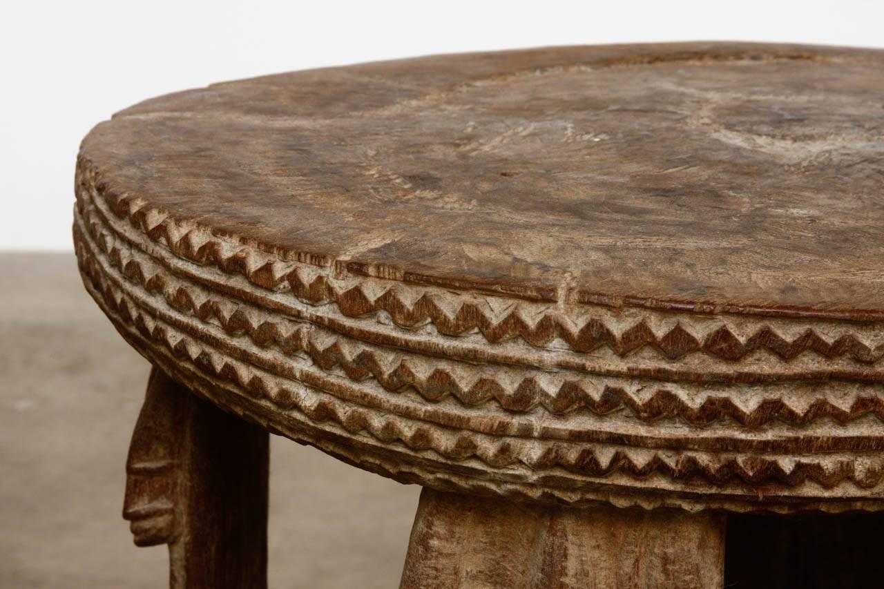 Primitive African carved stool or drinks table featuring carved figural supports. The top edge and base have decorative geometric patterns. The table is heavy and solid carved from one massive timber.