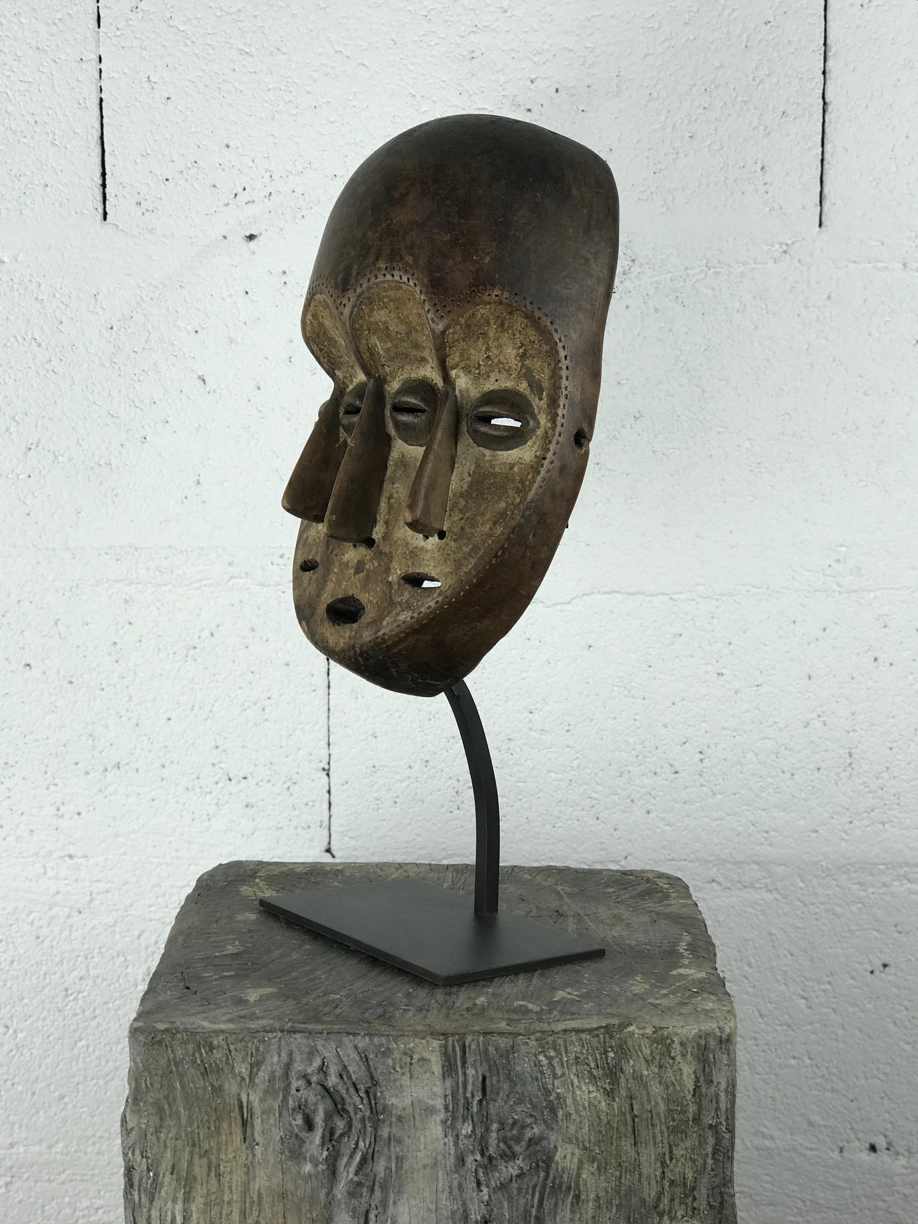A striking traditional African mask carved in wood, attributed to the Lega tribe of the Democratic Republic of Congo. This rare example features three stylized faces sharing one head, symbolizing wisdom, multiplicity of vision, and the ancestral
