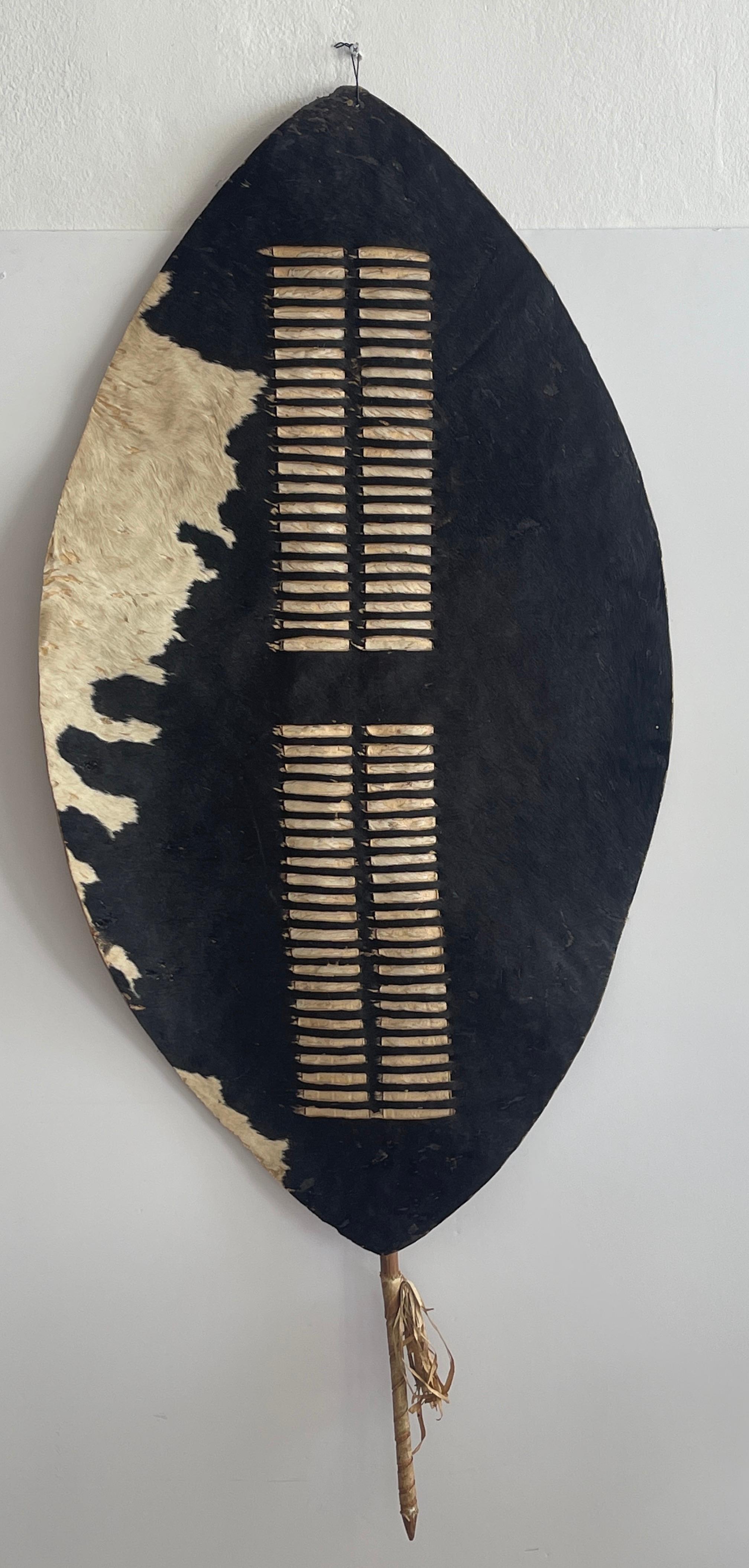 African Zulu cowhide shield, South Africa
Of oval form the cow hide shield with front with interwoven hairy strips, complete with pole at the back in form of a spear, with extending handle. 
Overall measurements 54” high x 23