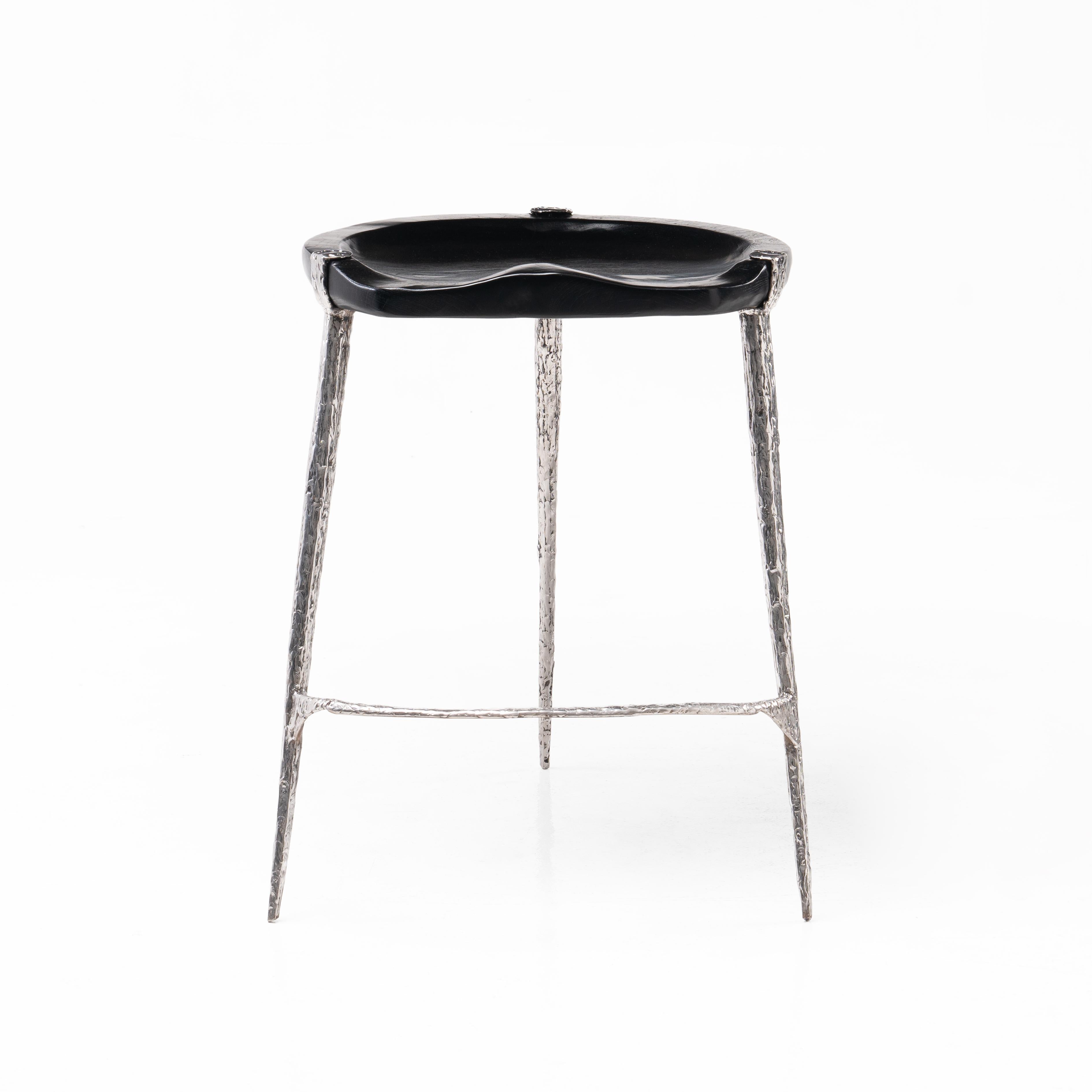 Ce tabouret sculptural présente des fragments d'acier inoxydable forgés à la main et fusionnés pour créer une esthétique organique qui reflète les textures trouvées dans les formations végétales. L'assise sculptée est réalisée à la main et présente