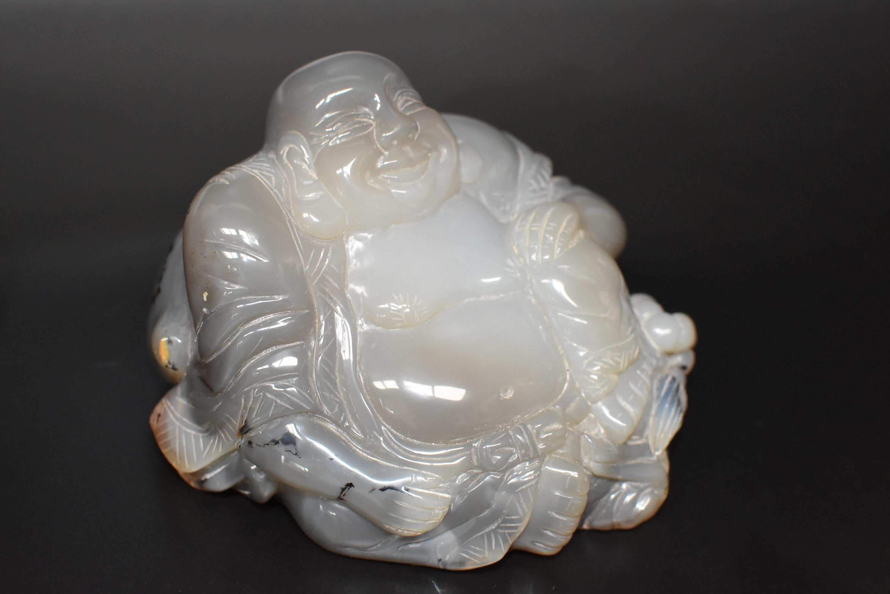 Ein handgeschnitzter, massiver Achat-Buddha. Er wird sitzend mit offenem Gewand dargestellt, das unter dem Bauch mit einer Schleife zusammengehalten wird, ein Ausdruck für große Zuwendung. Der glückliche Buddha ist eine geliebte Gottheit, die Glück