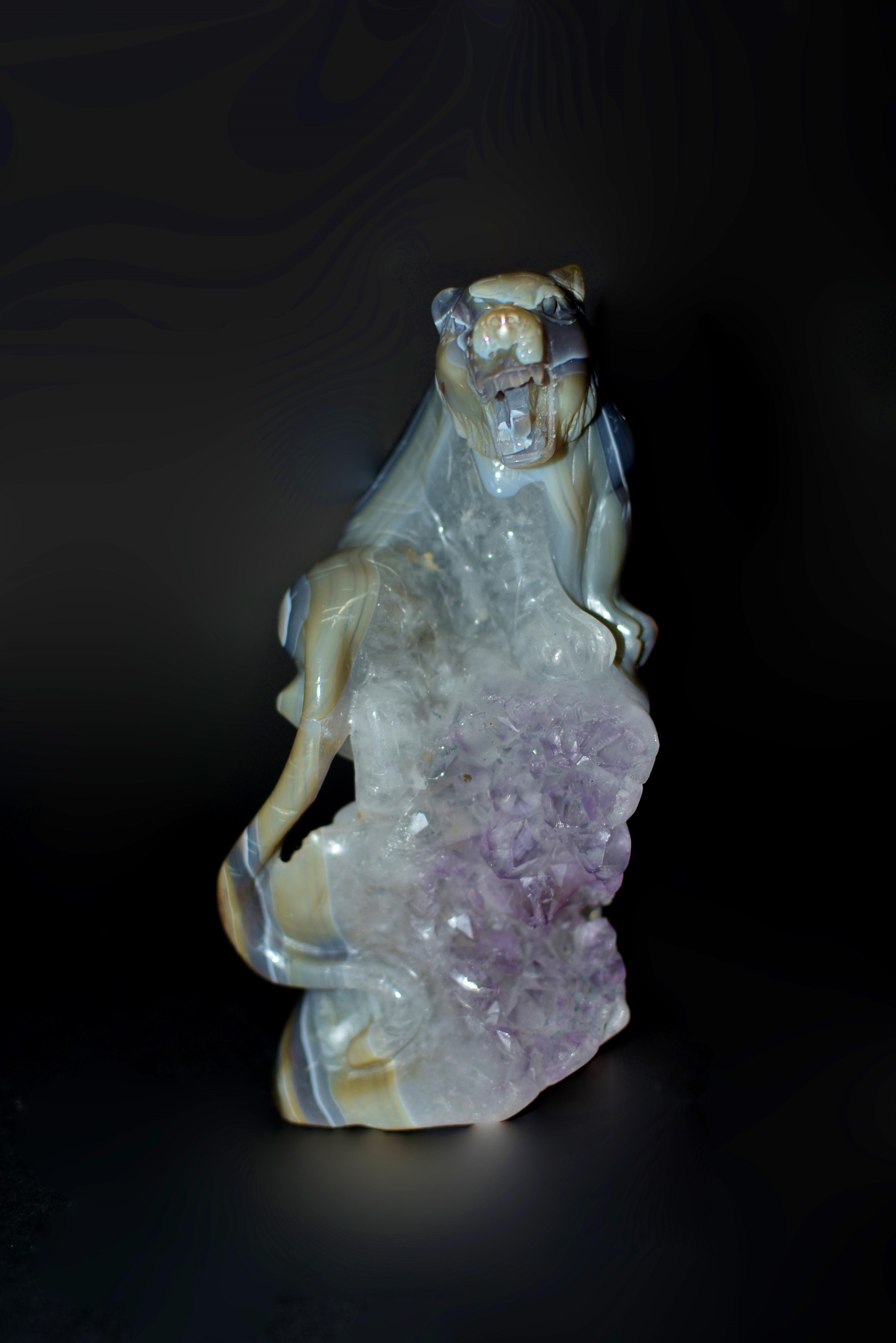 A beautiful Agate tiger on Amethyst rock sculpture, capturing the essence of raw power and untamed majesty. Poised upon a rocky outcrop of splendid purple amethyst, the tiger commands attention with its fierce gaze and open mouth, revealing menacing
