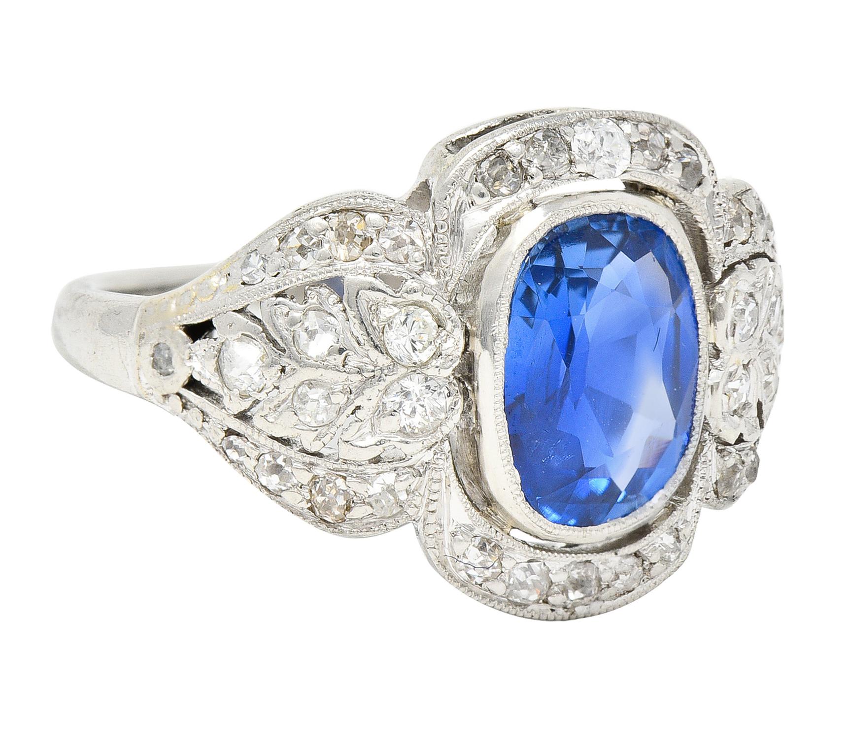 Centering a cushion cut sapphire weighing approximately 2.08 carats - transparent medium blue. Natural Burmese in origin with no indications of heat treatment - bezel set. Featuring a pierced foliate festoon motif surround. Bead and flush set with