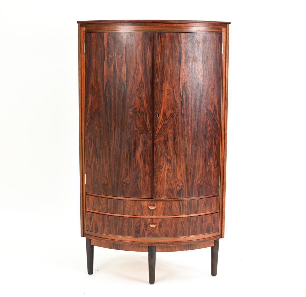 A gorgeous Danish mid-century round-front corner cabinet designed by Agner Christoffersen, c. 1950's. In handsome rosewood with an intricate bookmatched veneer. Featuring an upper cabinet with shelves and two drawers underneath. No manufacturer's