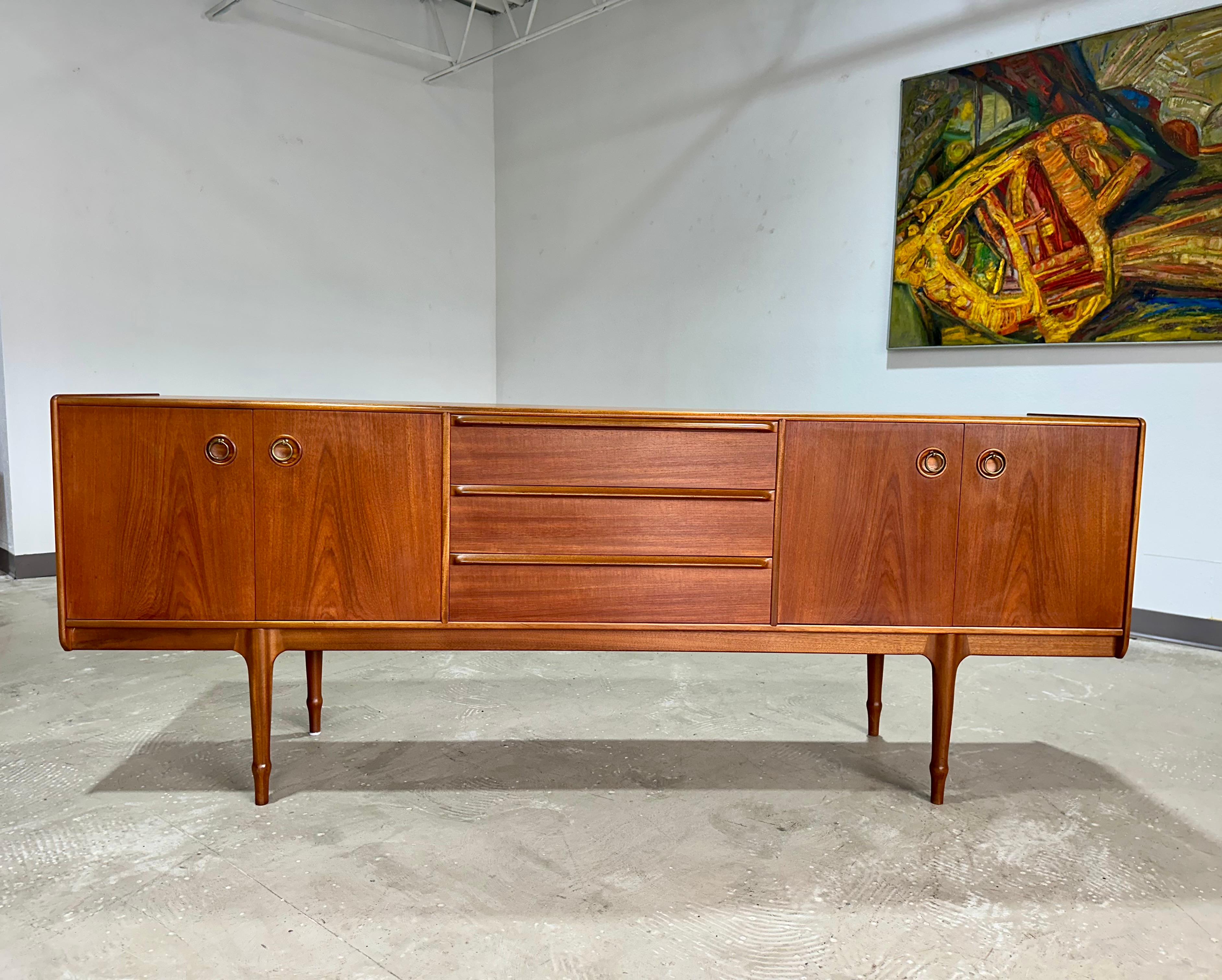 Vintage 1960’s Mid Century Modern extra long Teak credenza or sideboard attributed to A.H. McIntosh of Scotland. The teak case rests on long sculptural teak legs, features sleek curved front edges and highlighted by the inset brass circle pulls. An