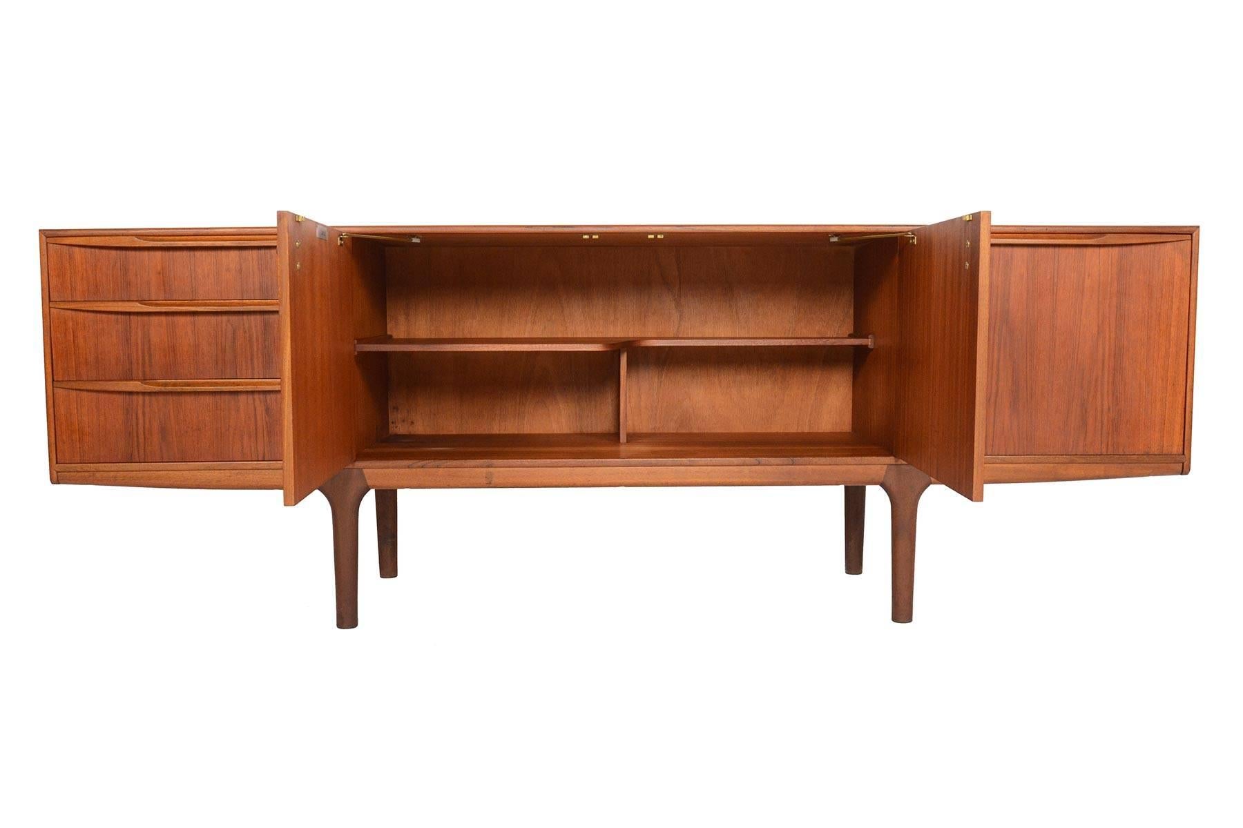 This Mid-Century Modern large low line teak sideboard by A.H. McIntosh was designed in the 1960s. Two centre doors open to reveal a fixed asymmetrical bar shelf. Right bar cabinet drops down to reveal open storage with a black laminate slide out