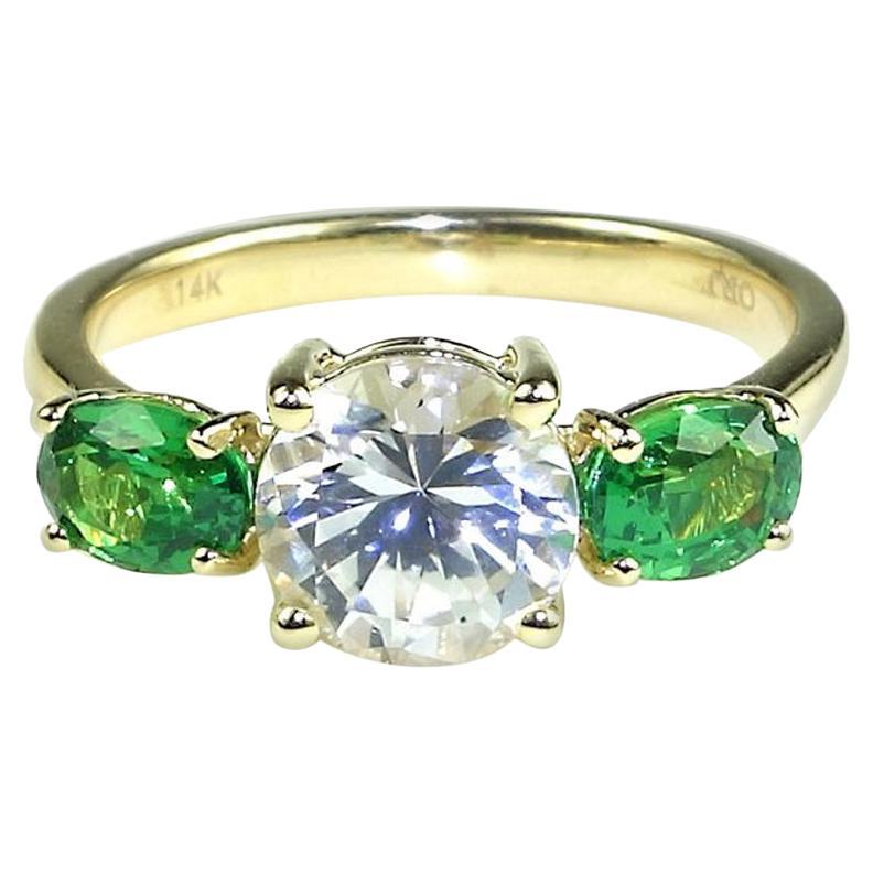 Dieser exquisite, maßgefertigte Ring aus 14 Karat Gelbgold ist ein atemberaubendes Symbol für Romantik und Eleganz. In der Mitte des Rings befindet sich ein glänzender, runder weißer Saphir, der durch zwei ovale grüne Tsavoriten ergänzt wird. Der