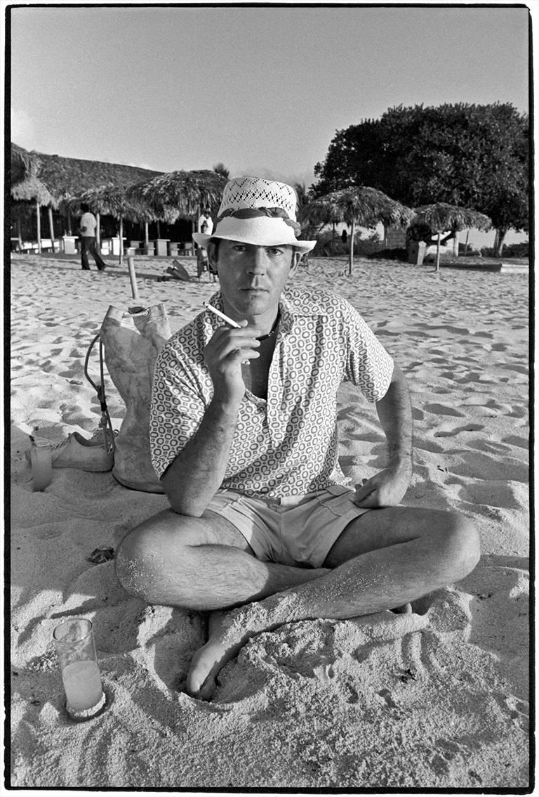 Hunter S. Thompson von Al Satterwhite zeigt ein Porträt von Hunter S. Thompson, der mit einer Zigarette in der Hand und einem Drink in der Hand am Strand sitzt. 

Dieses Foto ist als 20 x 16 Zoll großer Archivpigmentdruck in einer Auflage von 25