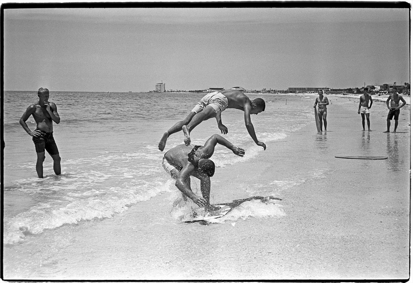 Surf Skimmers von Al Satterwhite fängt den unbeschwerten und fröhlichen Geist von Jugendlichen ein, die am Strand spielen,  Ein Junge springt über einen anderen und taucht dabei fast kopfüber in den Sand, während eine Gruppe in der Ferne zusieht.