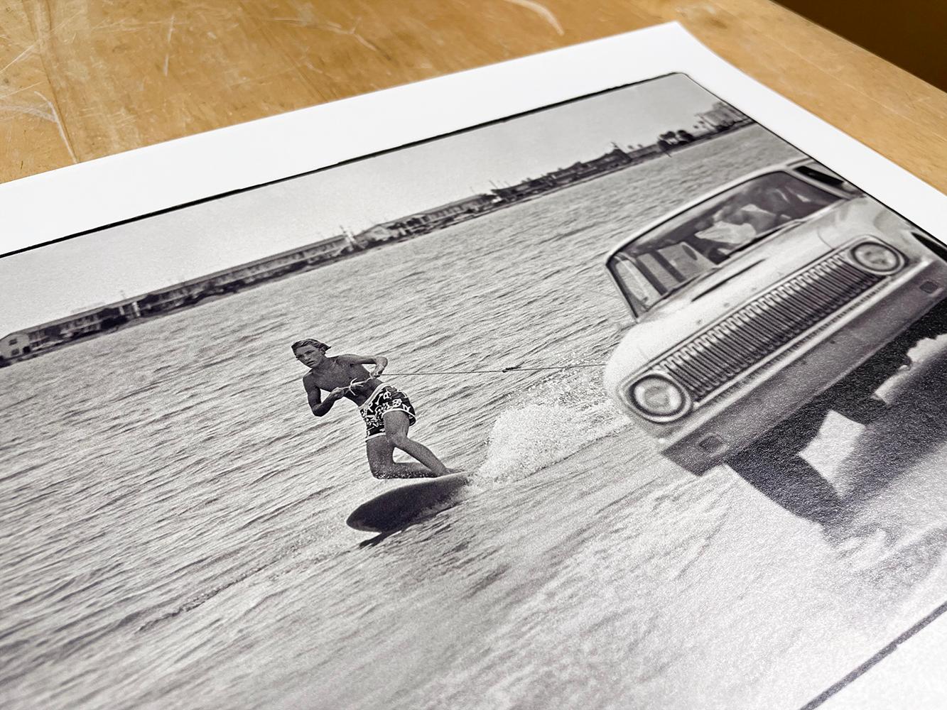 Surfing Florida Style von Al Satterwhite zeigt einen jungen Mann beim Surfen auf dem Wasser. Er hält sich an einem Seil fest, das an einem Auto befestigt ist, das ihn durch das Wasser zieht, während das Auto am Ufer entlang fährt. 

Archivalischer