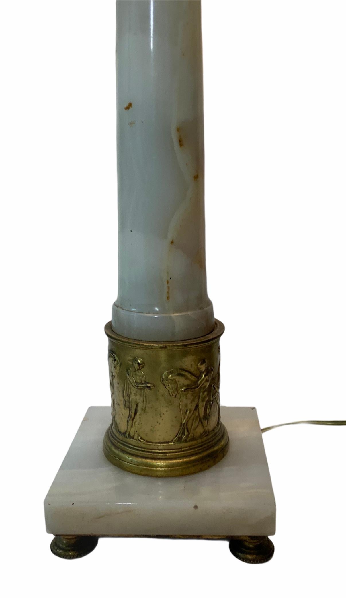 A translucent white alabaster with brown “veins” lamp build as an ascending column adorned with a bronze carved shell urn vase in the top and at the bottom a repousse bronze scene of Roman soldiers training horses. It stands in a square alabaster