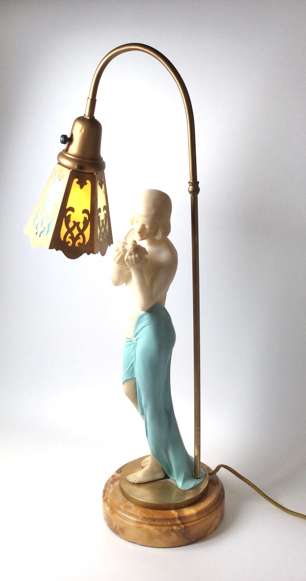 Jolie lampe figurative représentant une jolie femme portant un drapé bleu couvrant sa hanche, semi-nue sentant un bouquet de roses. Abat-jour de forme hexagonale avec 3 panneaux en verre de scorie de couleur bleue et crème. Elle repose sur un disque