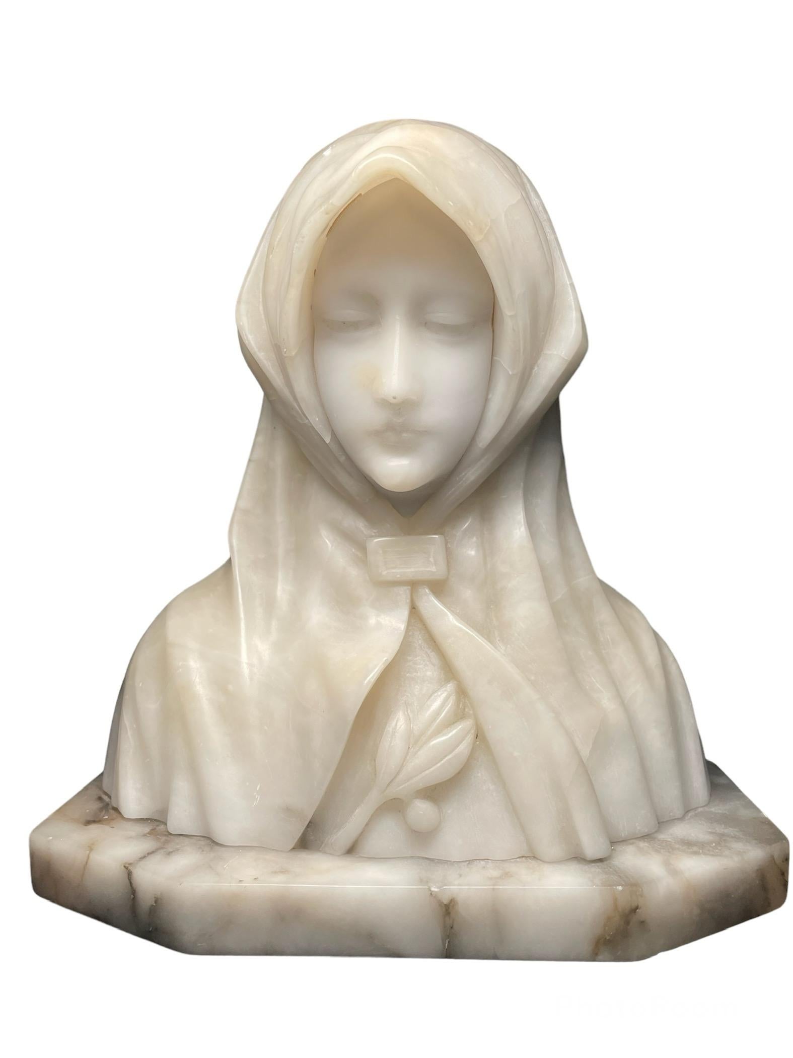 This is an alabaster and marble bust of Saint Clare of Assisi. It depicts a young lady head/face covered with a cloak. She is looking down, very peacefully while she appears to be praying to God. There is a branch of olive that are adorning her
