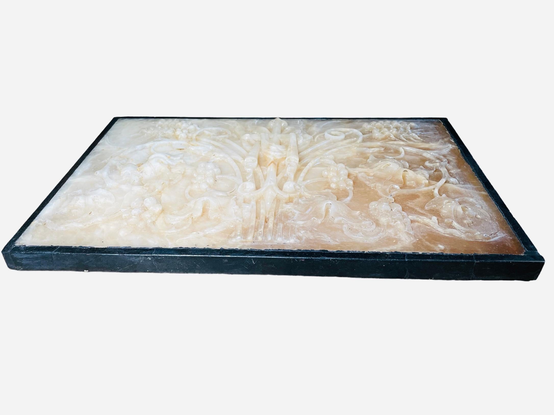 This an Alabaster and Marble Plaque. It depicts a long heavy hand carved alabaster plaque decorated with a cross in the center adorned with long scrolls of grapes vines leaves and wheat branches. Two wings adorn the sides of the upper top of the