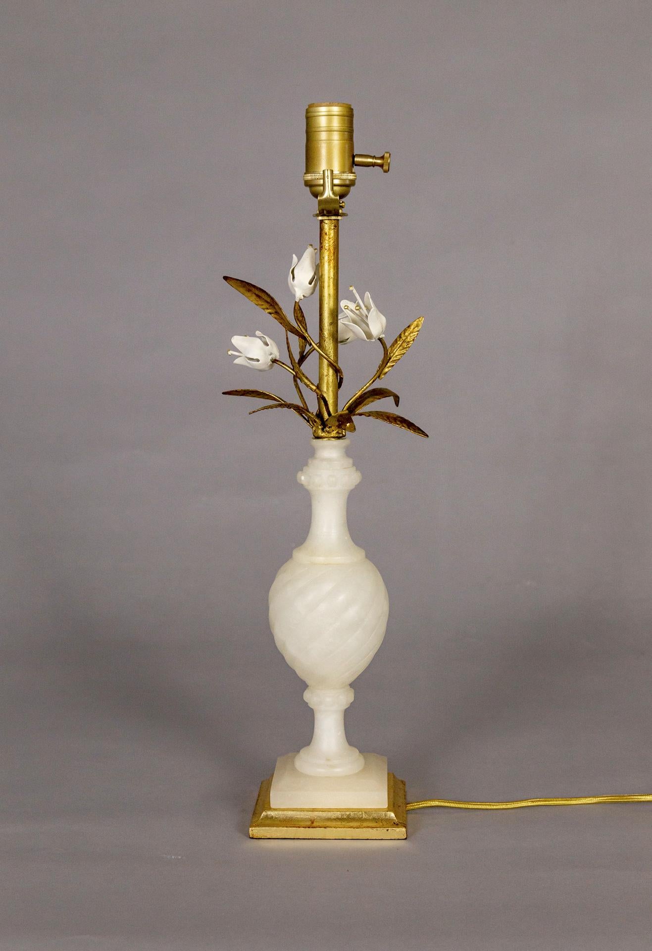 A newly restored table lamp of carved alabaster topped with white painted flowers and gilded metal leaves around the brass stem. It sits on a gilded, wood base. The stone is carved in a baluster shape with subtle, diagonal ribbing on the main body.