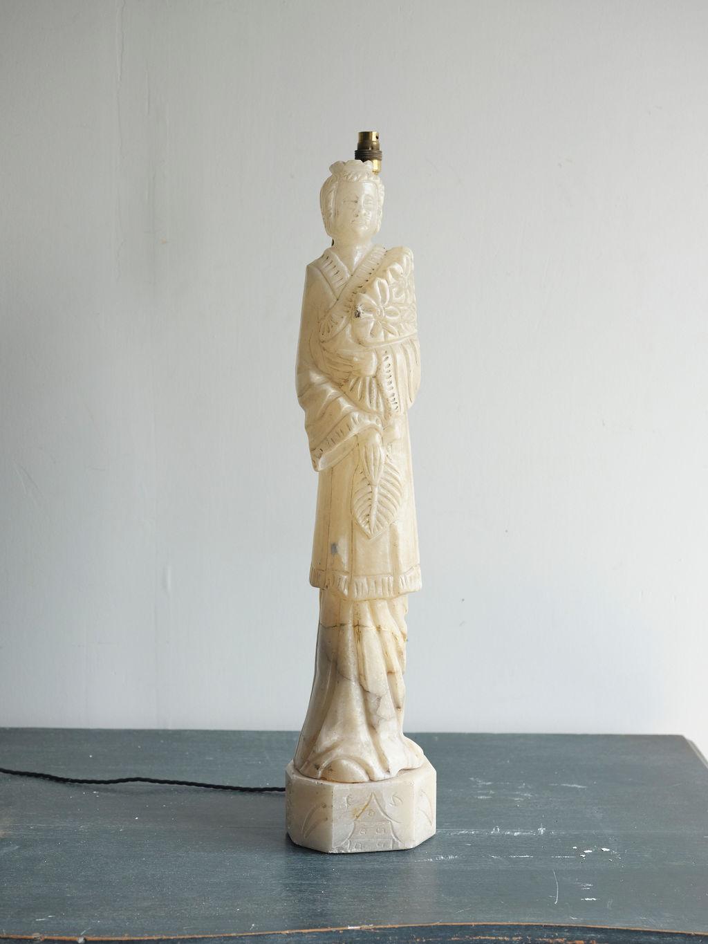This is a lovely figurative alabaster lamp, created in 1860. The lamp figure appears to be a woman in a decorative cloak, holding a leaf. The color is a creamy off-white color. This lamp is sturdy. It is European wired.