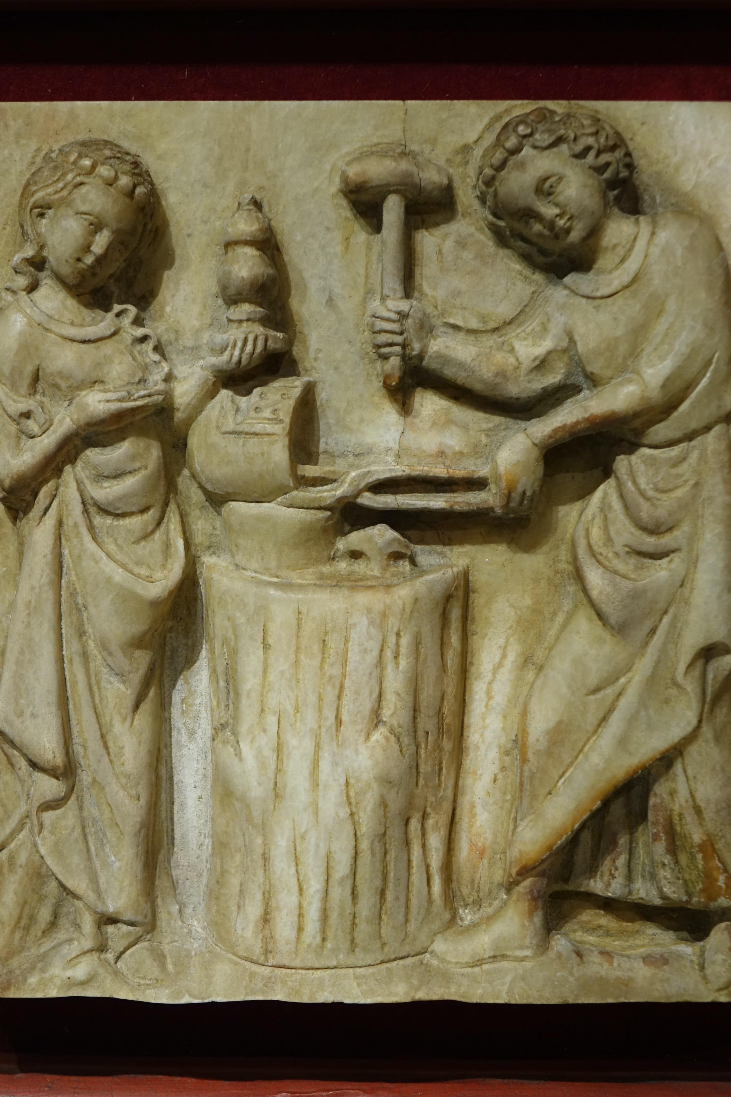 Alabaster plate in high relief illustrating a forgeron working on a metal object (a helmet?). A young woman is in front of him holding in her left hand an ovoid vase or reliquary that she is holding in her right hand.
The scene represented is being