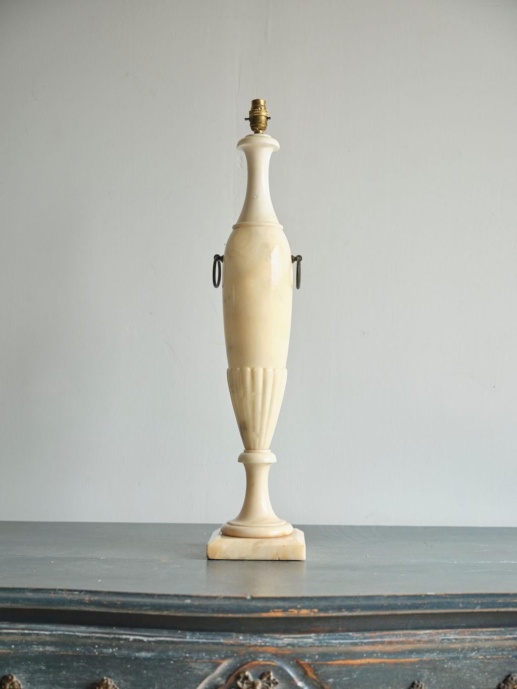This is a lovely 19th century alabaster lamp. It is a creamy color and has a unique shape. It is very sturdy. This lamp does not come with a lamp shade. It has two decorative brass rings on either side and stands on a square base. This lamp is