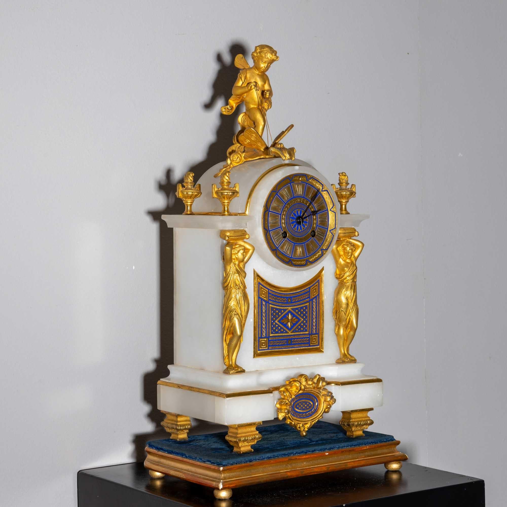 Pendule de cheminée dans un boîtier en albâtre à heures romaines, décor bleu de cobalt et doré en forme de cariatides, d'urnes enflammées et d'un putto ailé chevauchant un papillon. L'horloge est posée sur un piédestal recouvert de velours bleu.