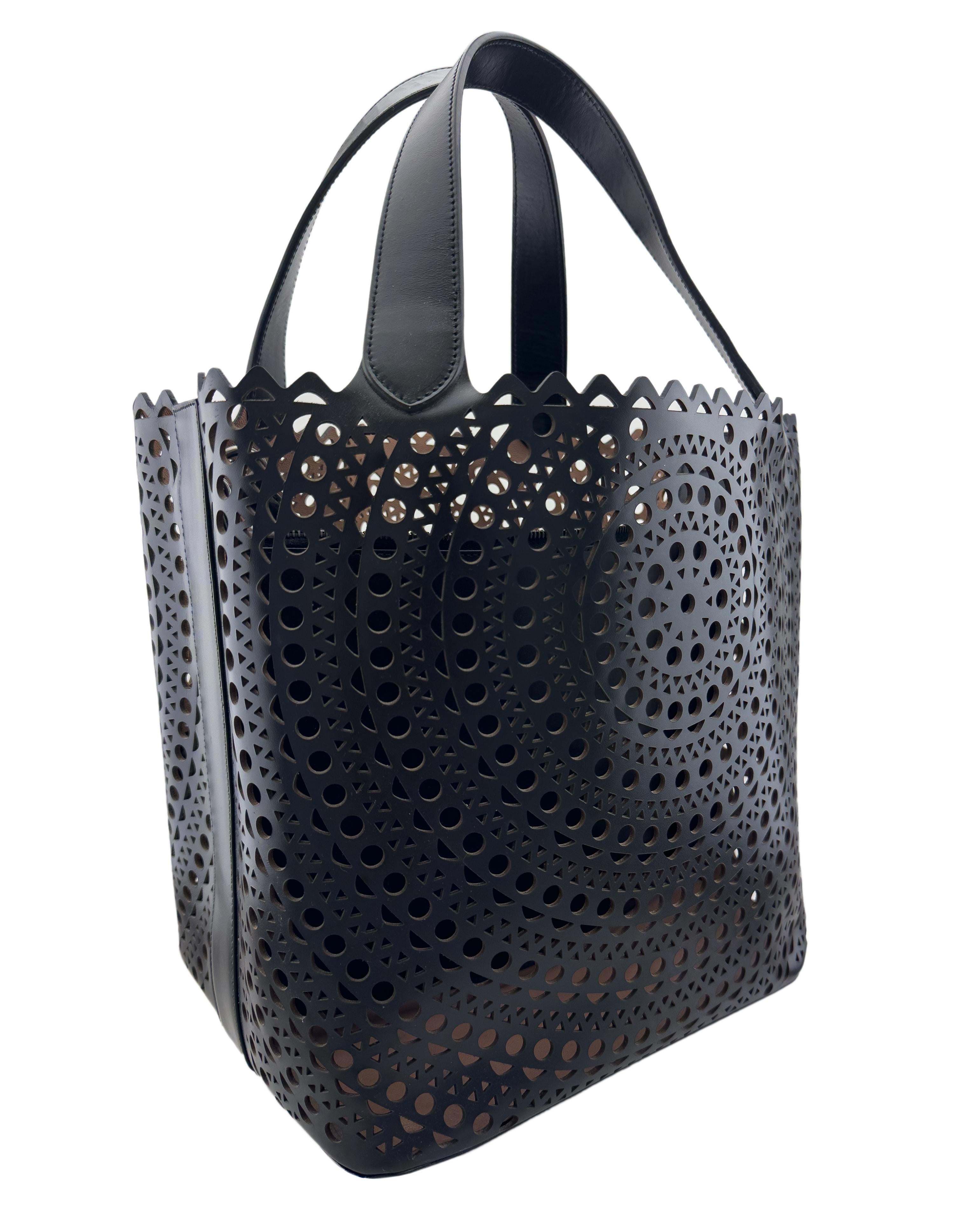 Alaia black tote crafted from leather that features scalloped and laser cut accents on the exterior. It has protective feet at base with engraved logo, an open-top that reveals a leather-lined interior with a single large interior pocket and enough