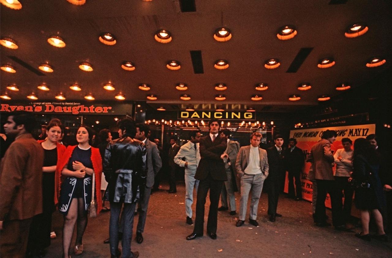 Waiting To Dance by Alain Le Garsmeur
Young people waiting at the entrance to the film and dancing arcade in Piccadilly, London, England, 1972.

Paper size 30 x 40 inches / 76 x 101 cm  
Printed in 2022 - produced from the original